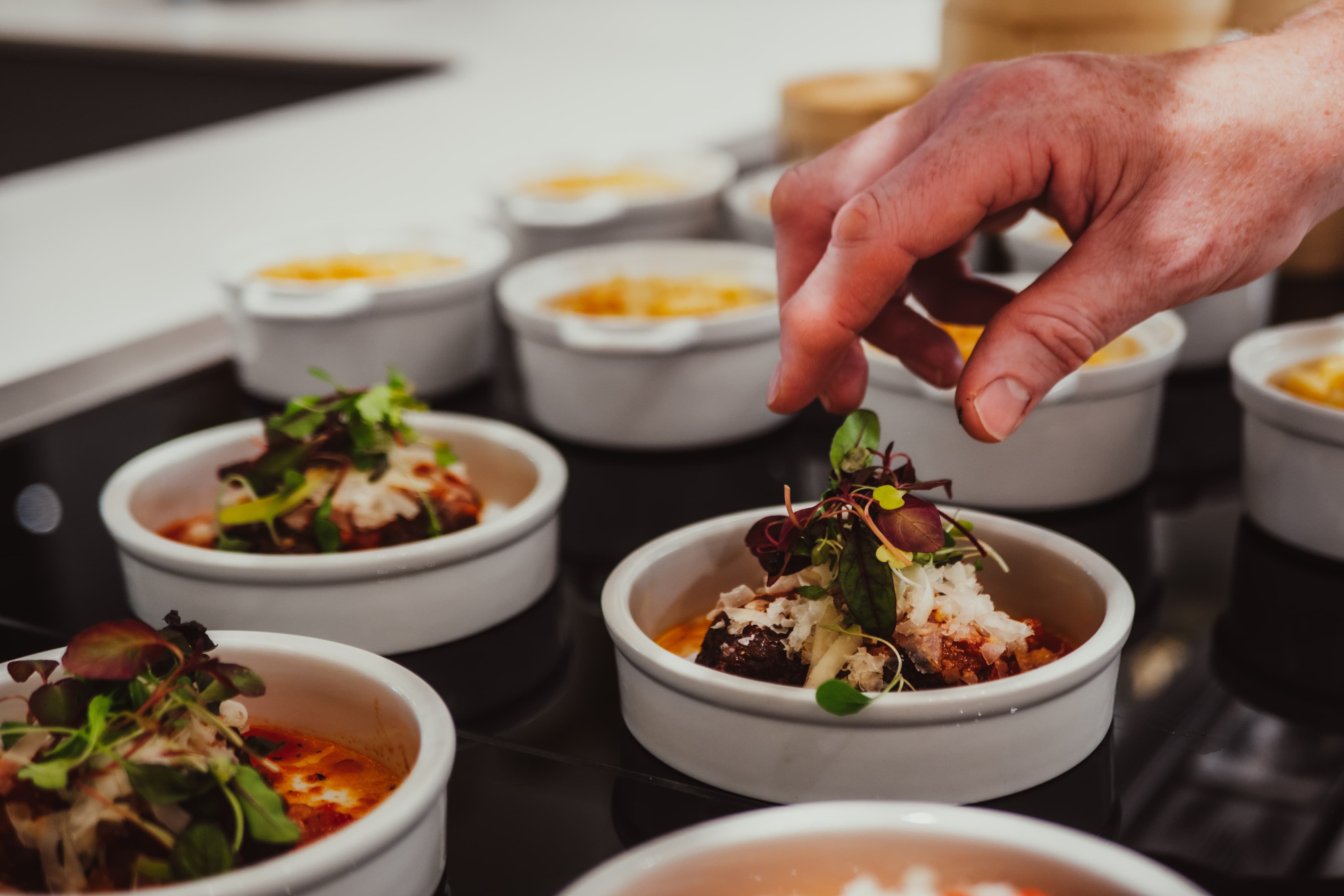 A hand garnishing dishes with microgreens in small white bowls, each containing a prepared meal topped with grated cheese.