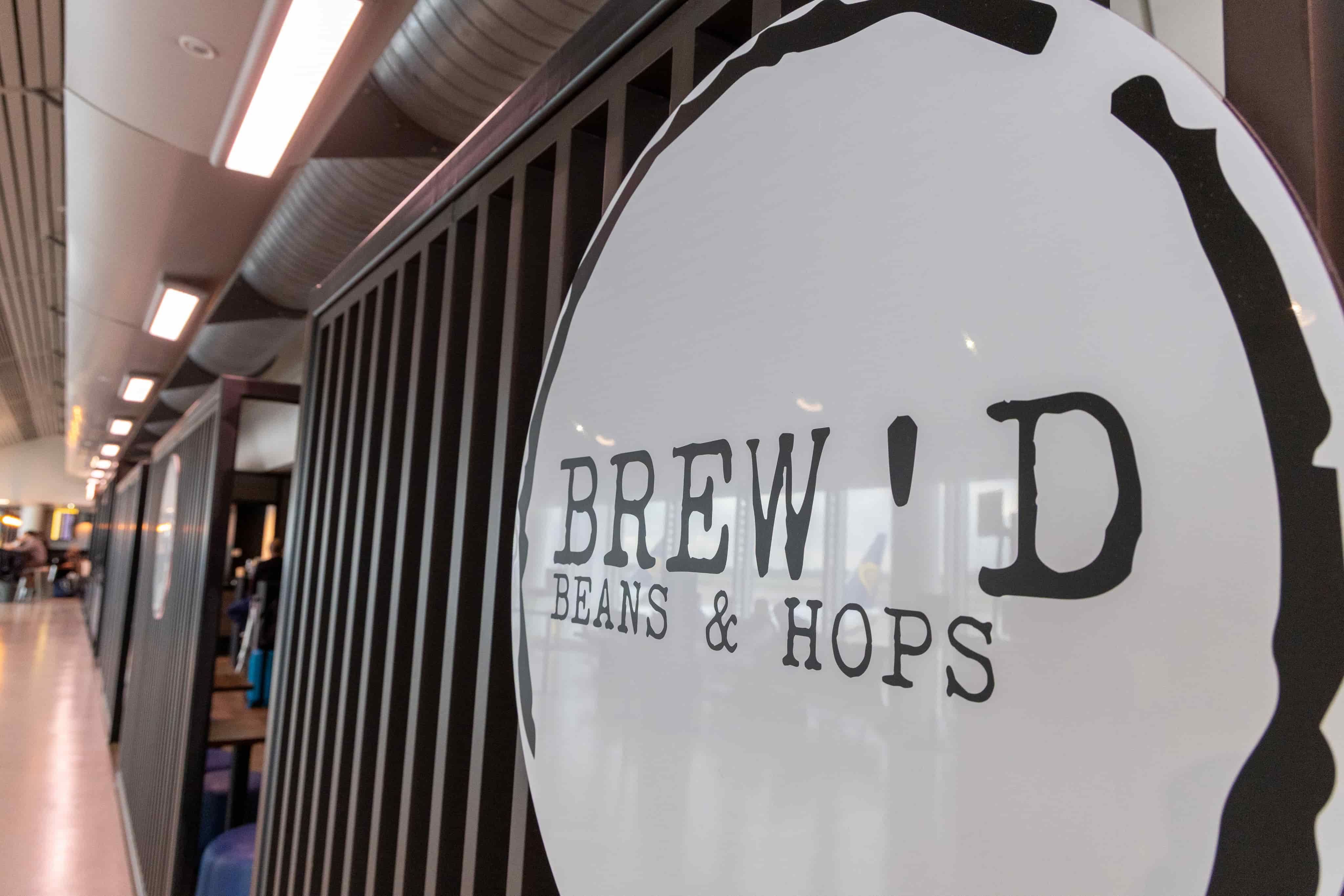 Sign reading "BREW'D BEANS & HOPS" on a wall with a modern interior and visible ceiling ductwork in the background.