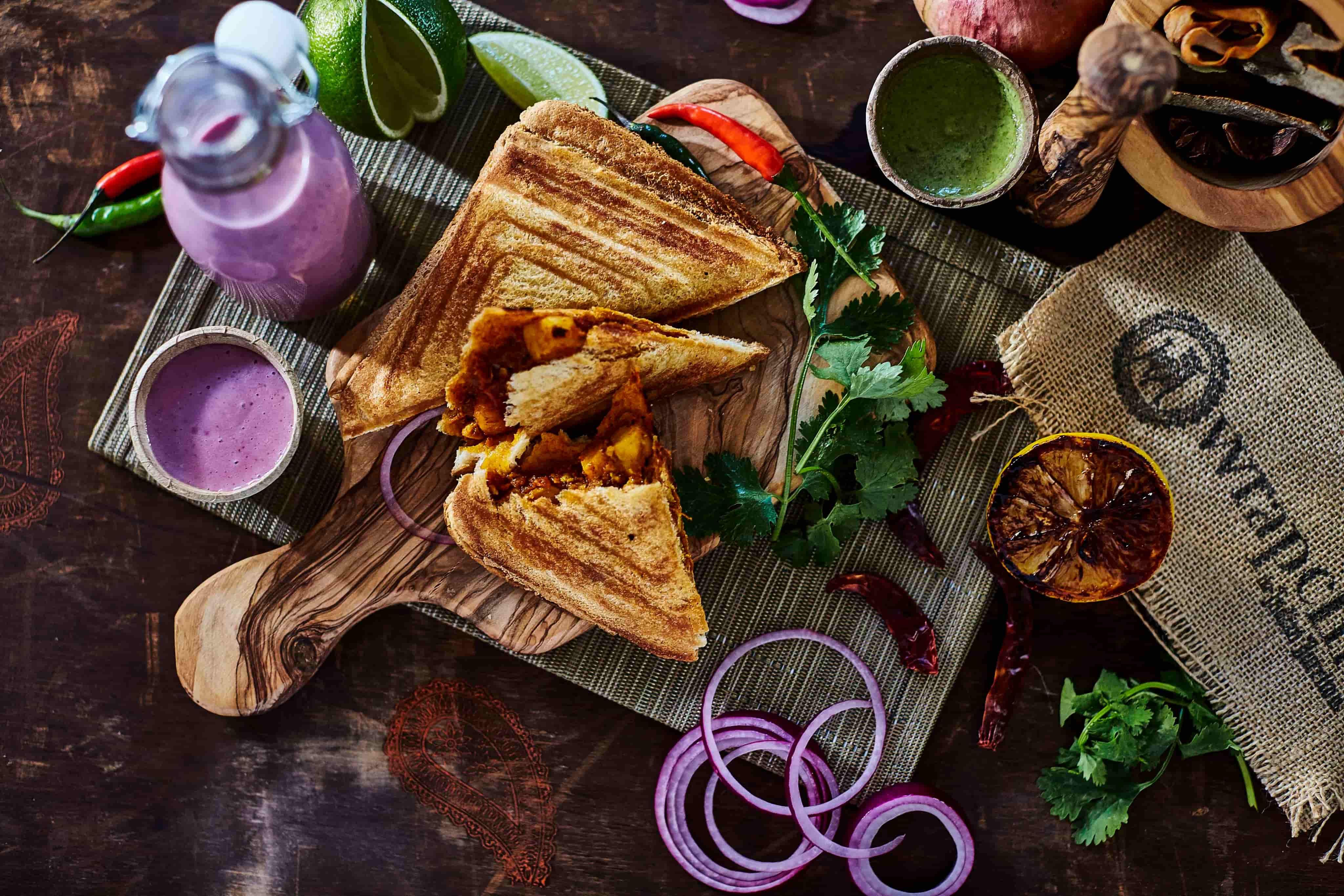 Toasted sandwiches with chutney on a wooden board, surrounded by sliced onions, herbs, and a bottle of pink beverage.