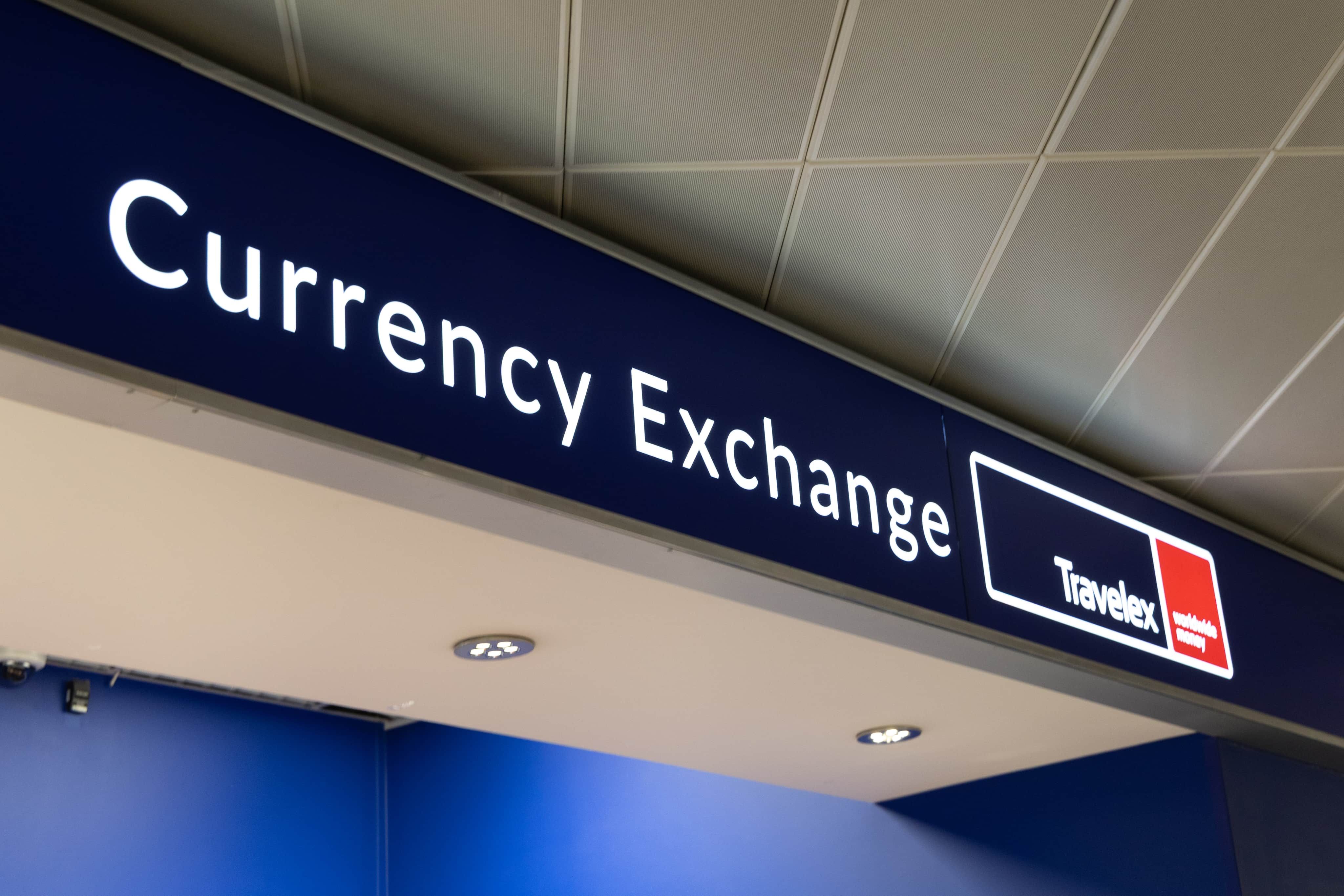 A sign for a currency exchange office with "Travelex" branding, mounted on the ceiling, under a grid-patterned tiled roof.