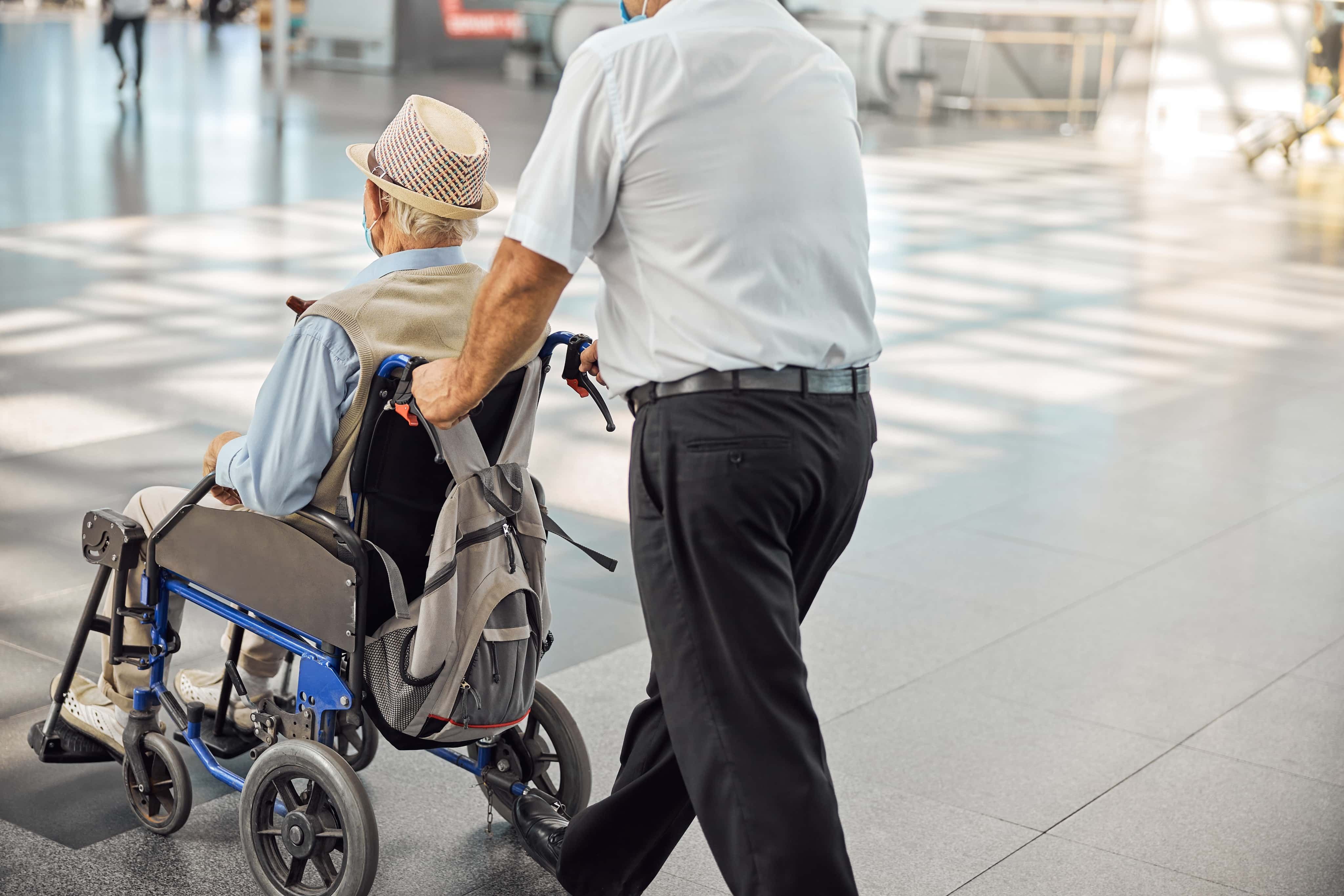 A person pushes an elderly man in a wheelchair through a spacious, well-lit area, with sunlight streaming through large windows.
