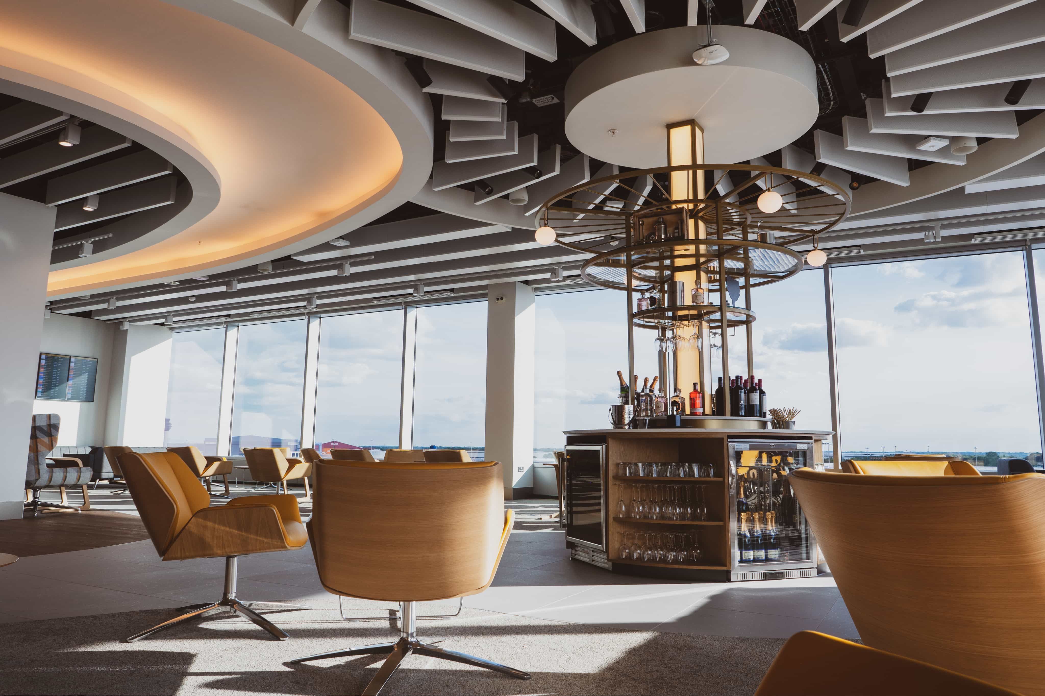 Modern lounge with circular bar, wooden chairs, and large windows offering a cityscape view. Unique ceiling design with ambient lighting.