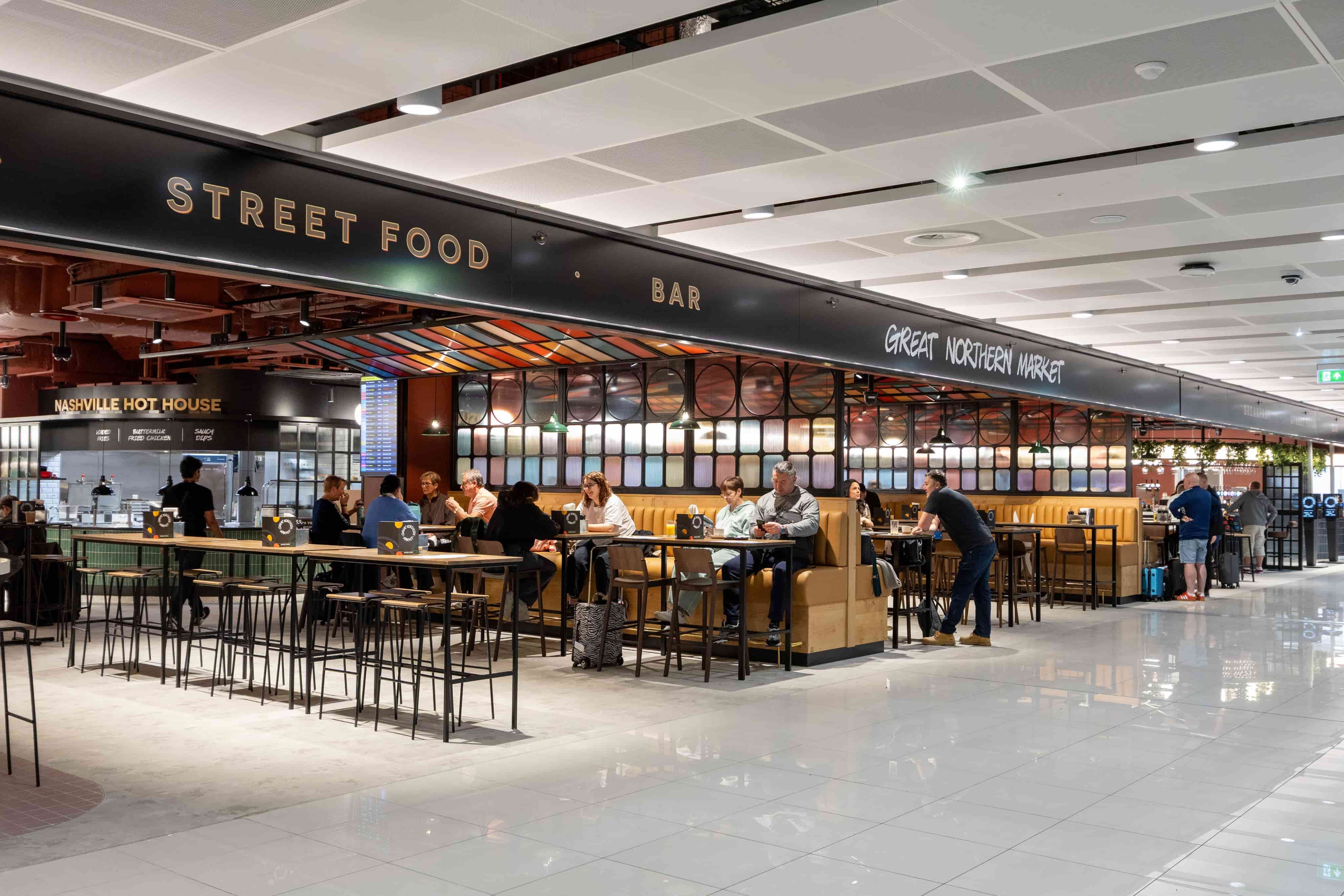 Modern airport food court with people dining at tables under a "Street Food" sign. Bright lighting and contemporary design.