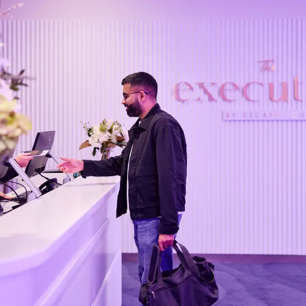 A man at a hotel reception counter checks in, handing over an ID while holding a bag. The background shows the logo "The Executive."