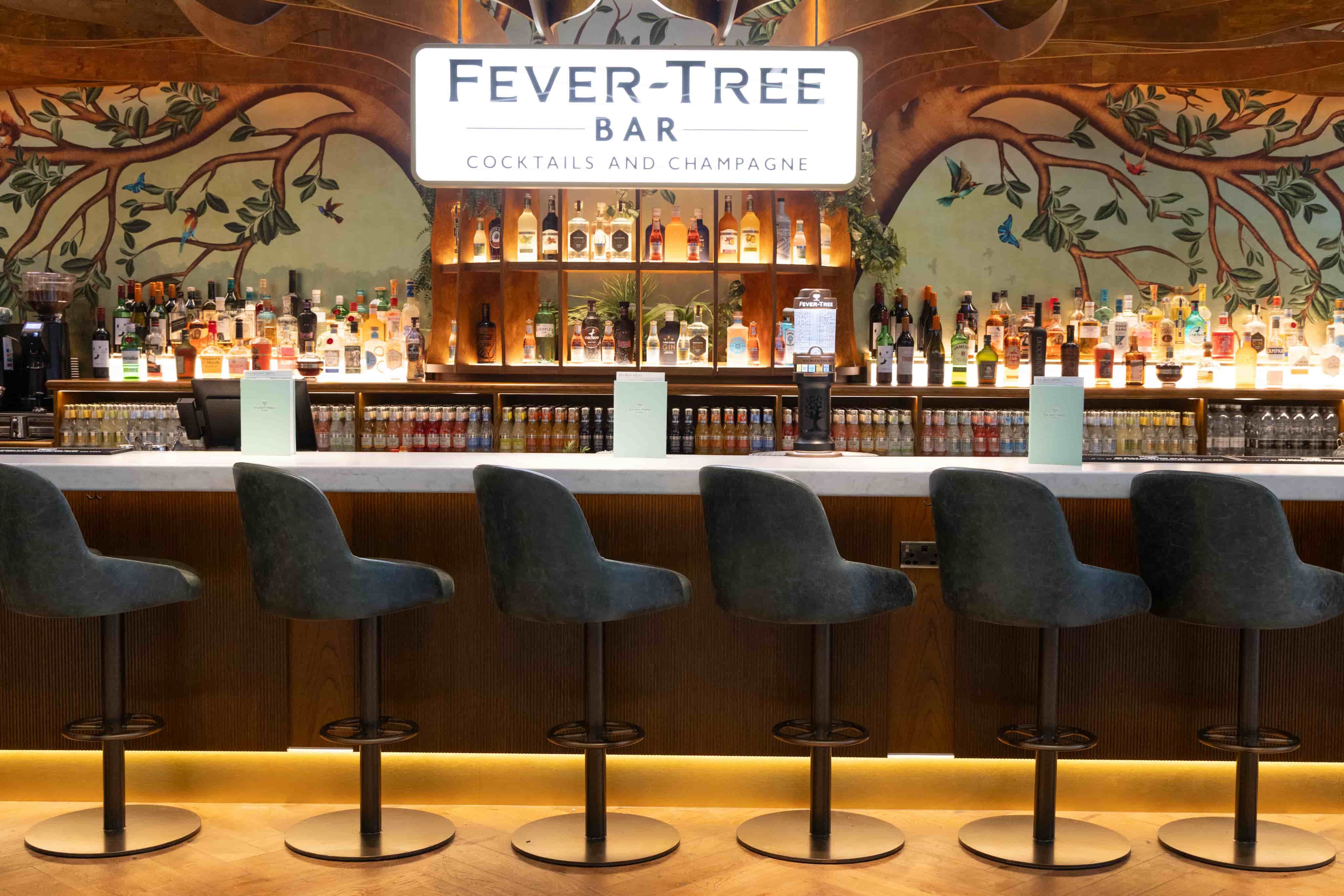 Modern bar with high stools facing a well-stocked liquor shelf. "Fever-Tree Bar" sign above, offering cocktails and champagne.