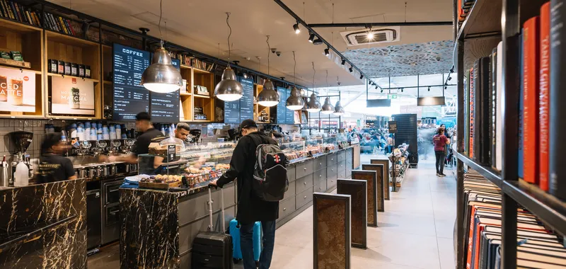 A cosy coffee shop with a marble counter, customers ordering drinks, hanging lights, and shelves filled with books and travel essentials.