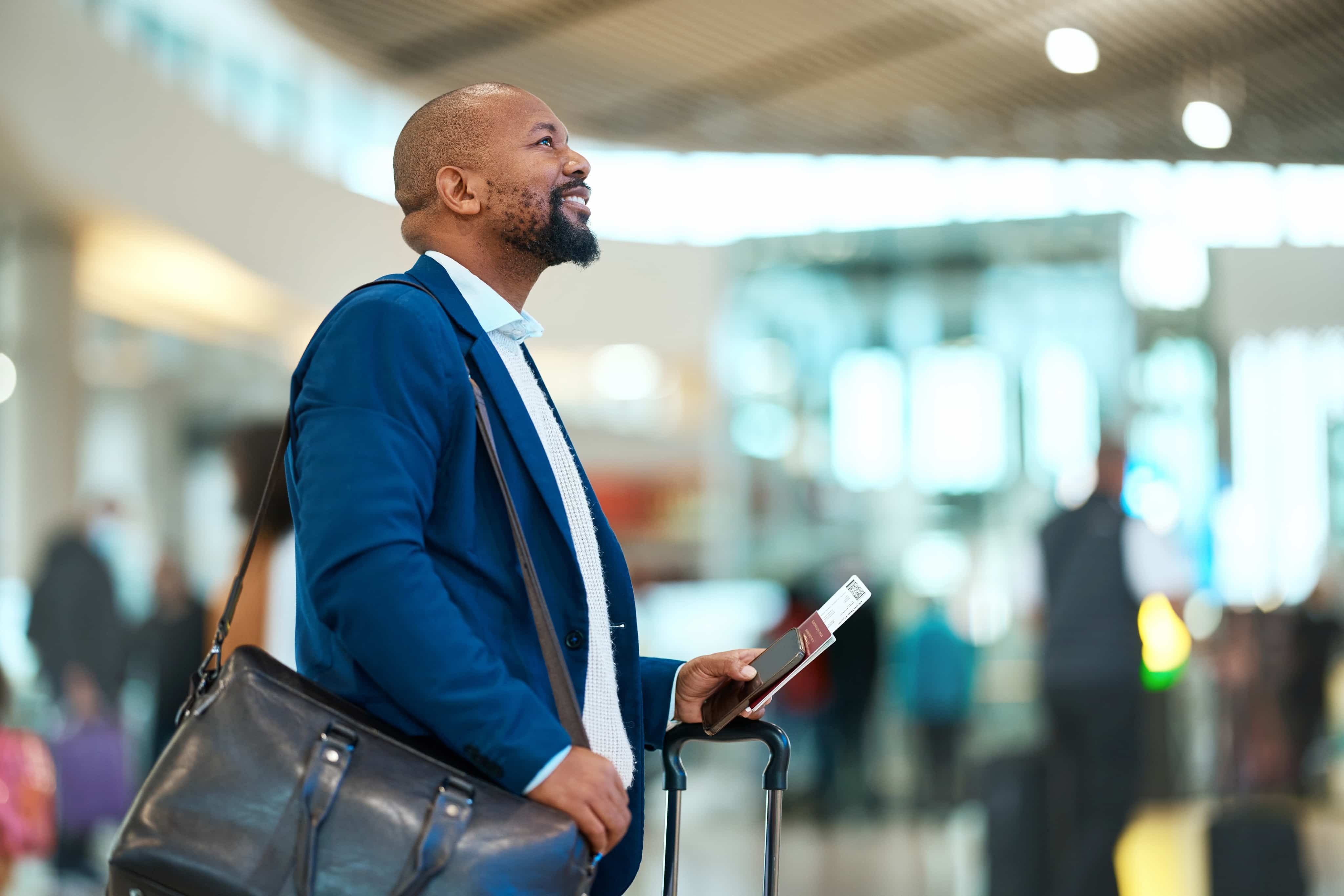 Man in a blue jacket holding a passport and suitcase, looking upwards in an airport terminal.