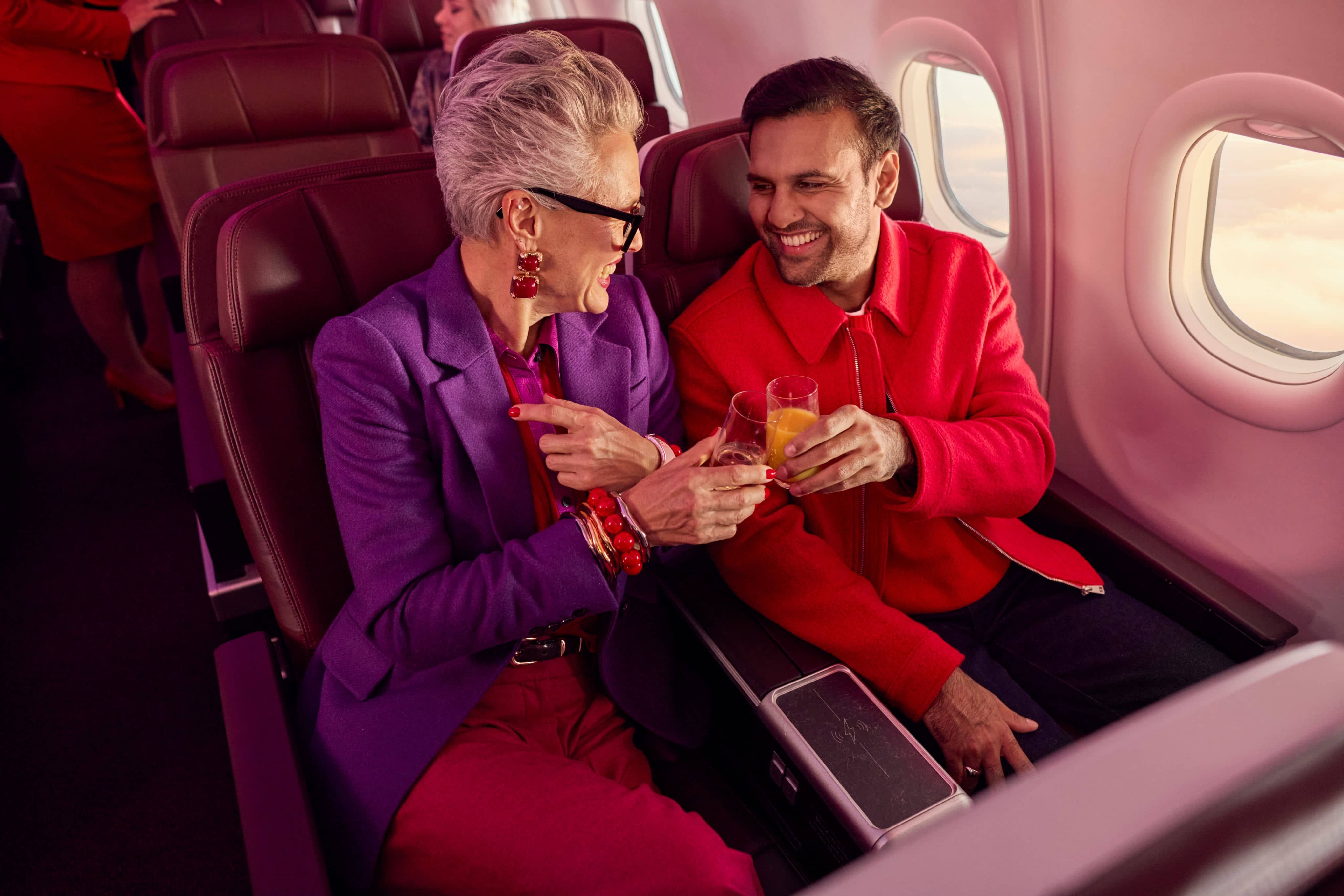 Two people seated on a plane, smiling and clinking drinks, against a view of clouds through the window.
