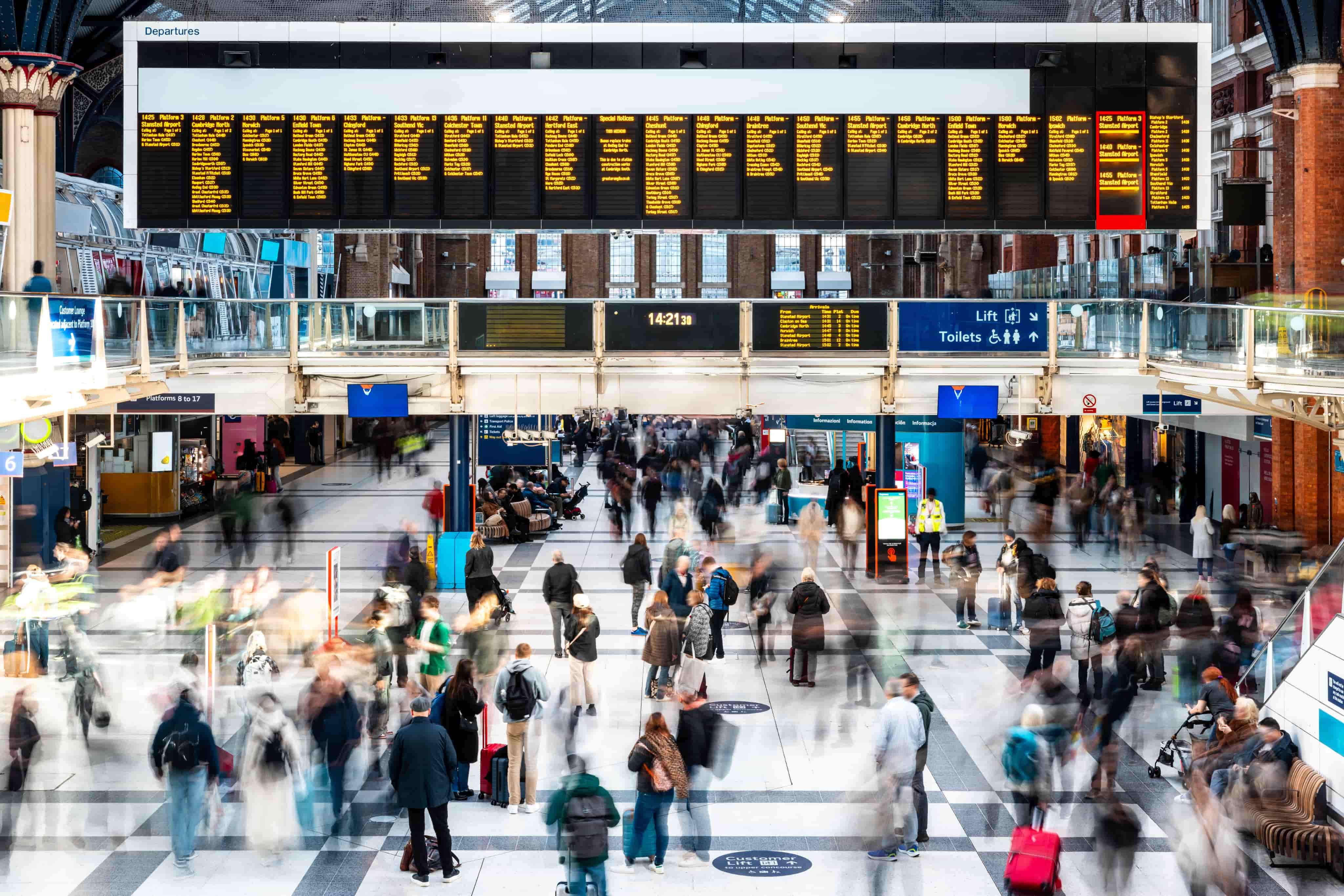 Busy train station with a large digital departure board and blurred motion of travelers walking and carrying luggage.