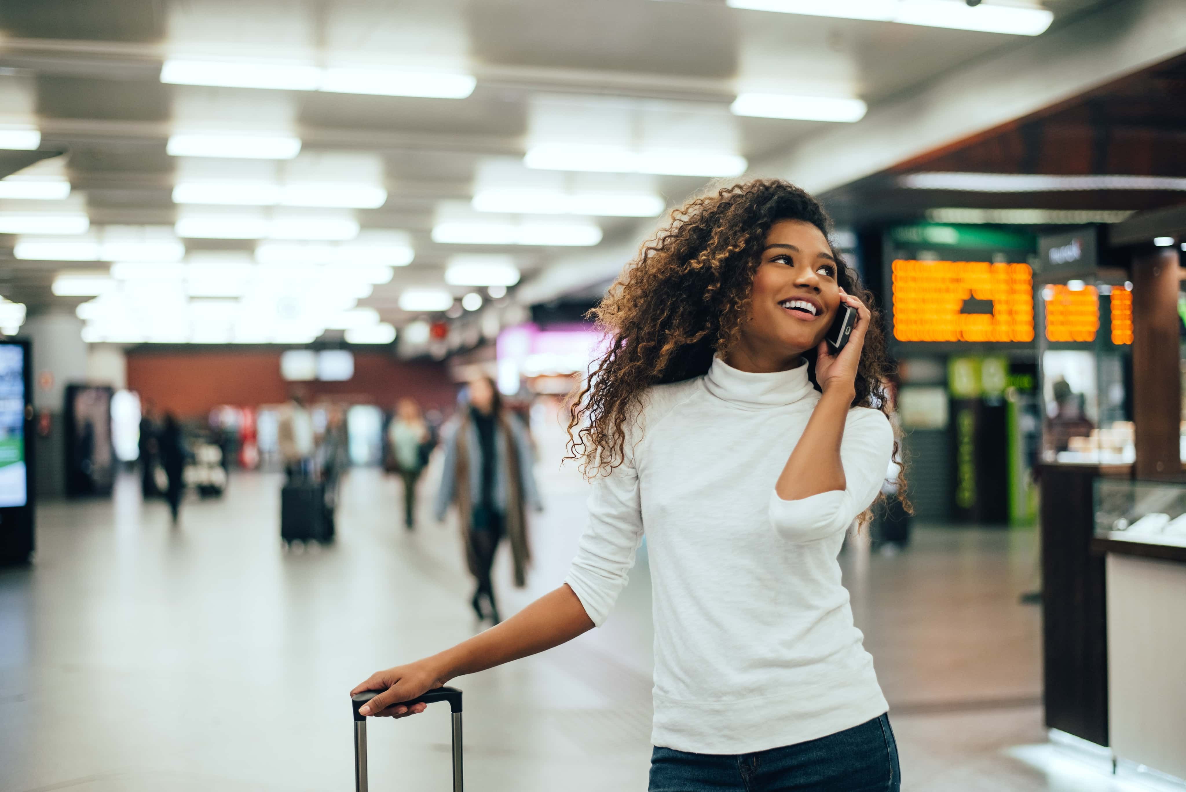 Woman with curly hair in a white sweater, smiling while talking on the phone, pulls a suitcase in a busy train station.