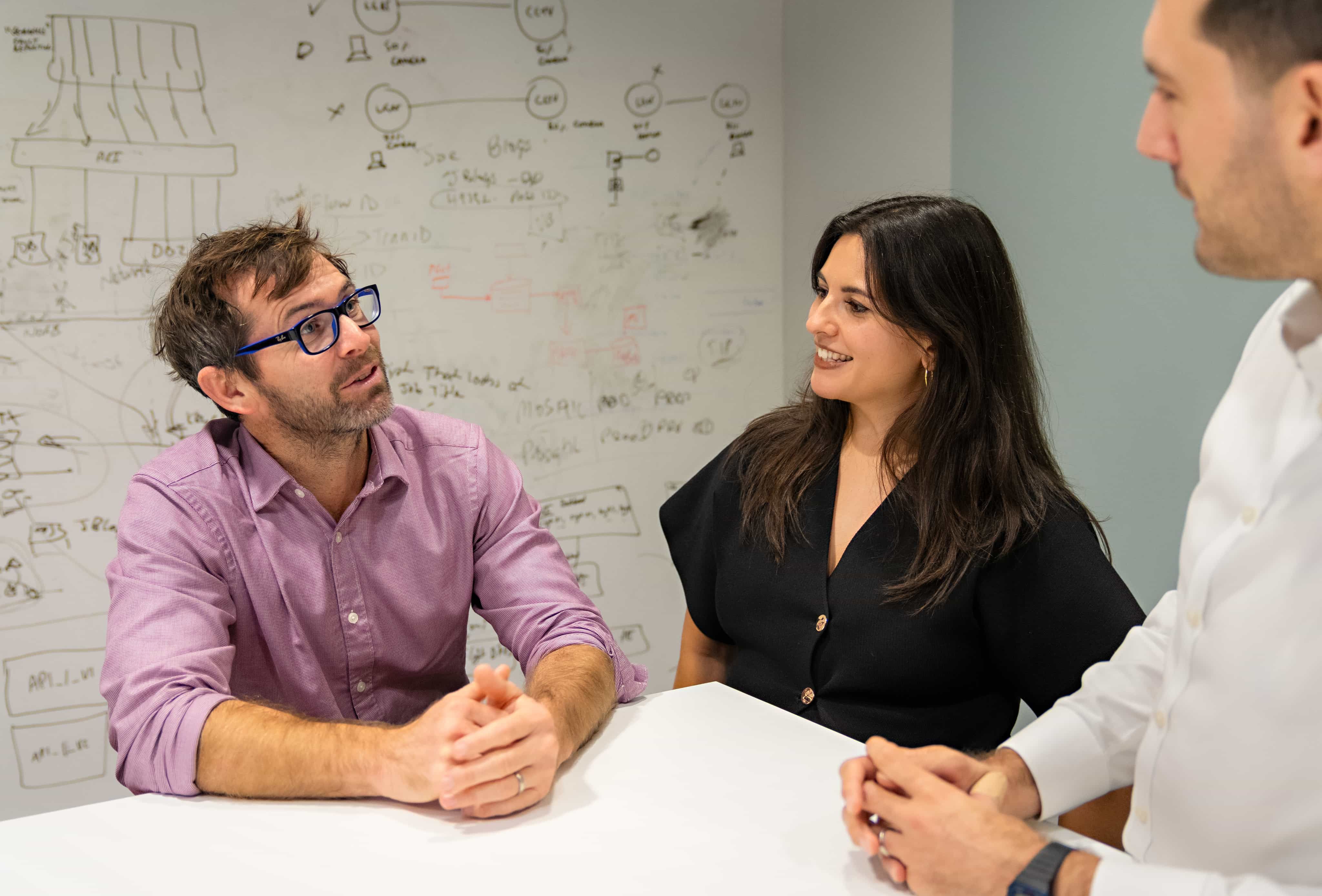 Three people discussing in a meeting room with a whiteboard covered in diagrams and notes in the background.