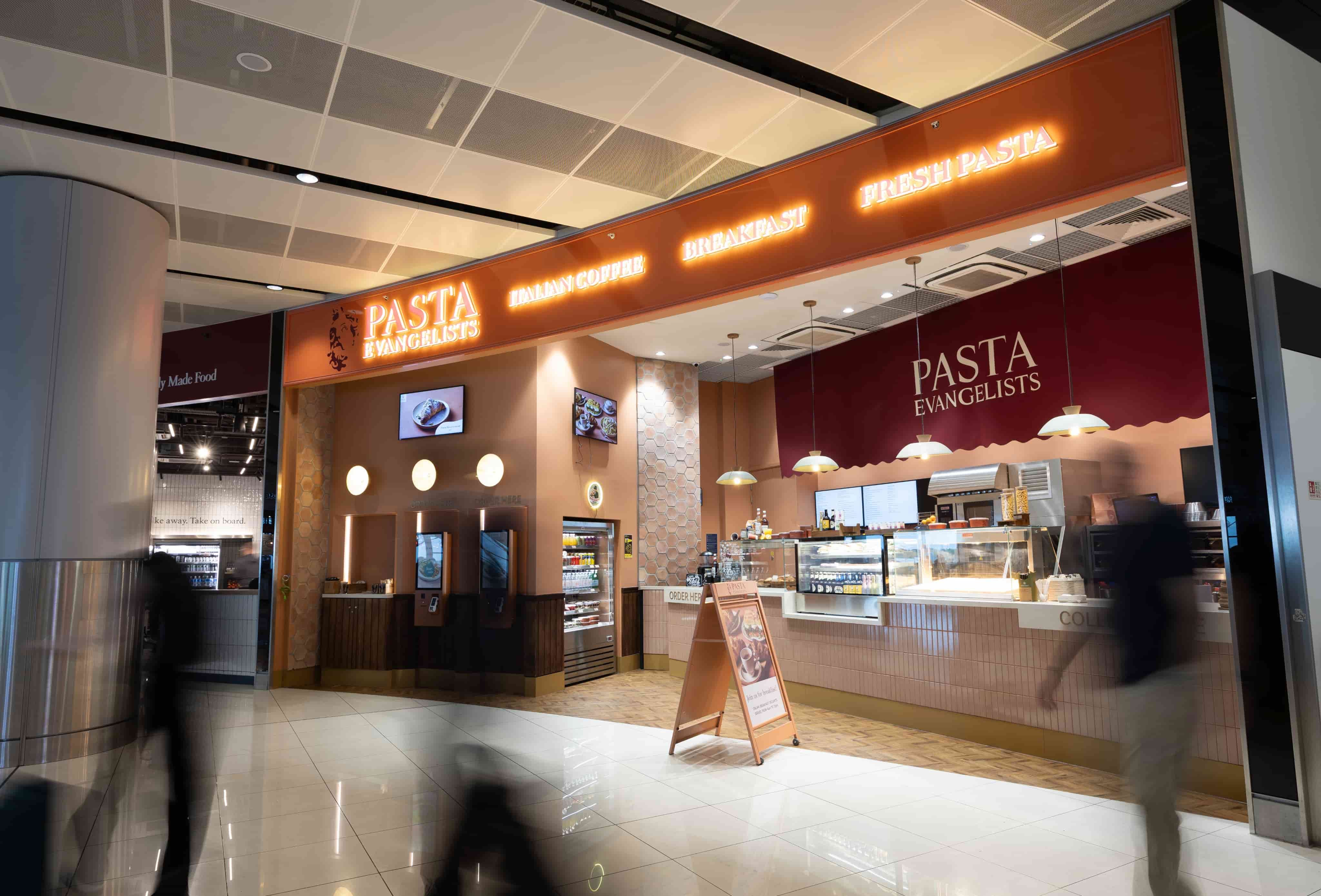 Pasta restaurant with bright orange signage in an indoor setting, featuring a counter, menu display, and blurred motion of passing people.