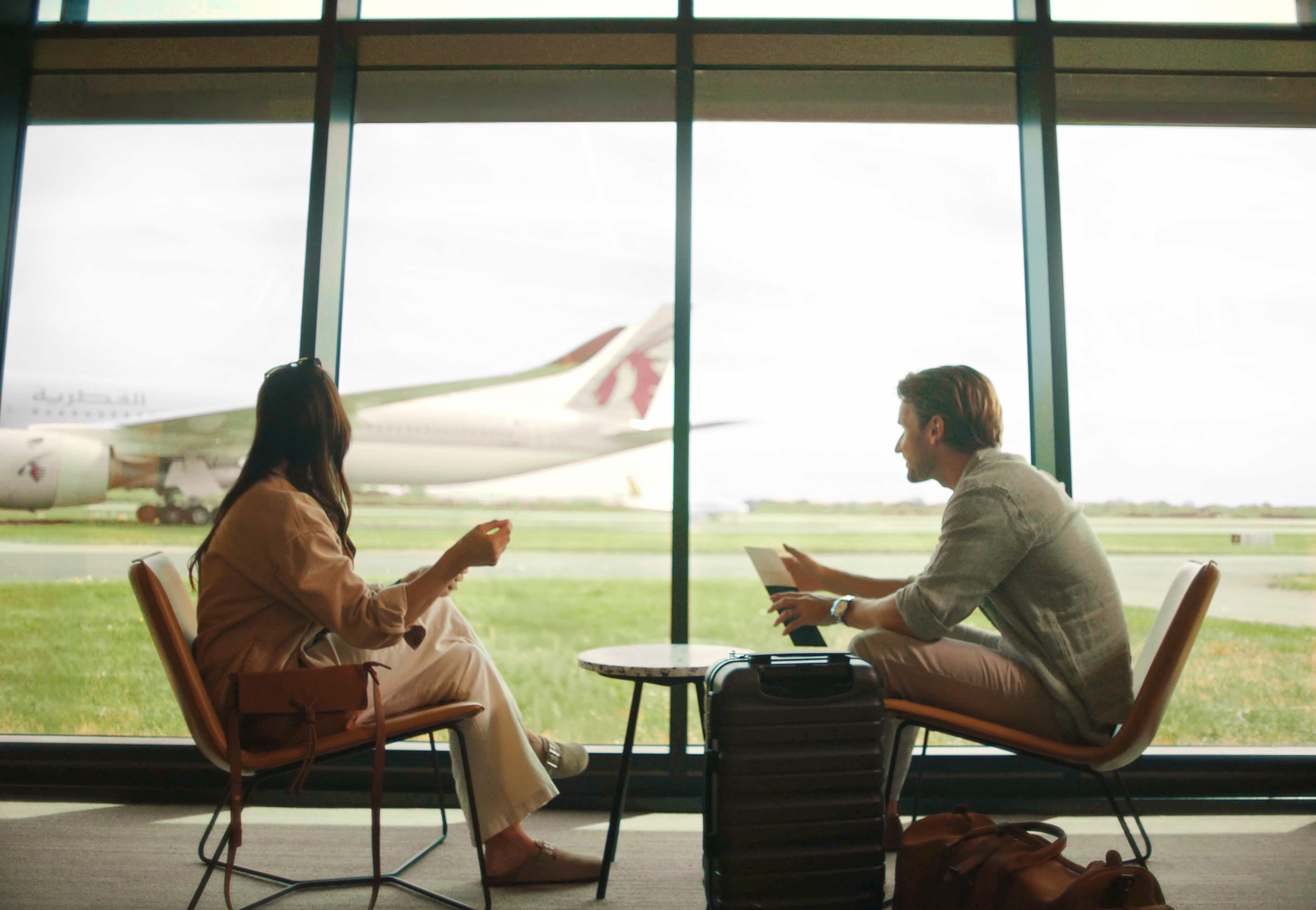 Two people sitting in an airport lounge, talking with a plane visible through large windows. A suitcase and bags are at their feet.