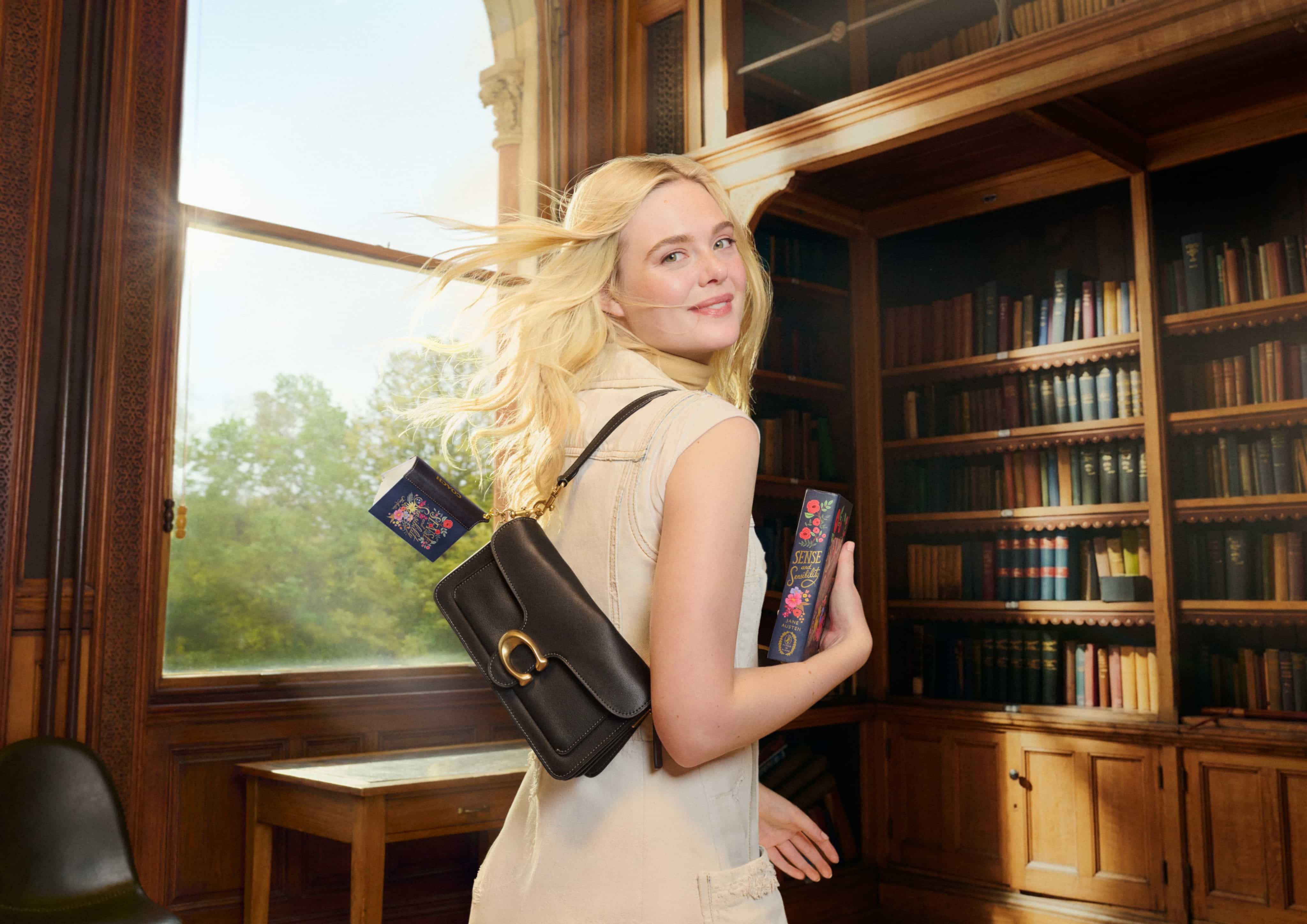 Woman with long blonde hair in a library, holding a book and wearing a brown shoulder bag, with sunlight streaming through a window.