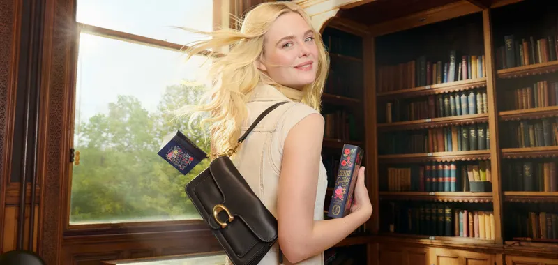 Woman with long blonde hair in a library, holding a book and wearing a brown shoulder bag, with sunlight streaming through a window.