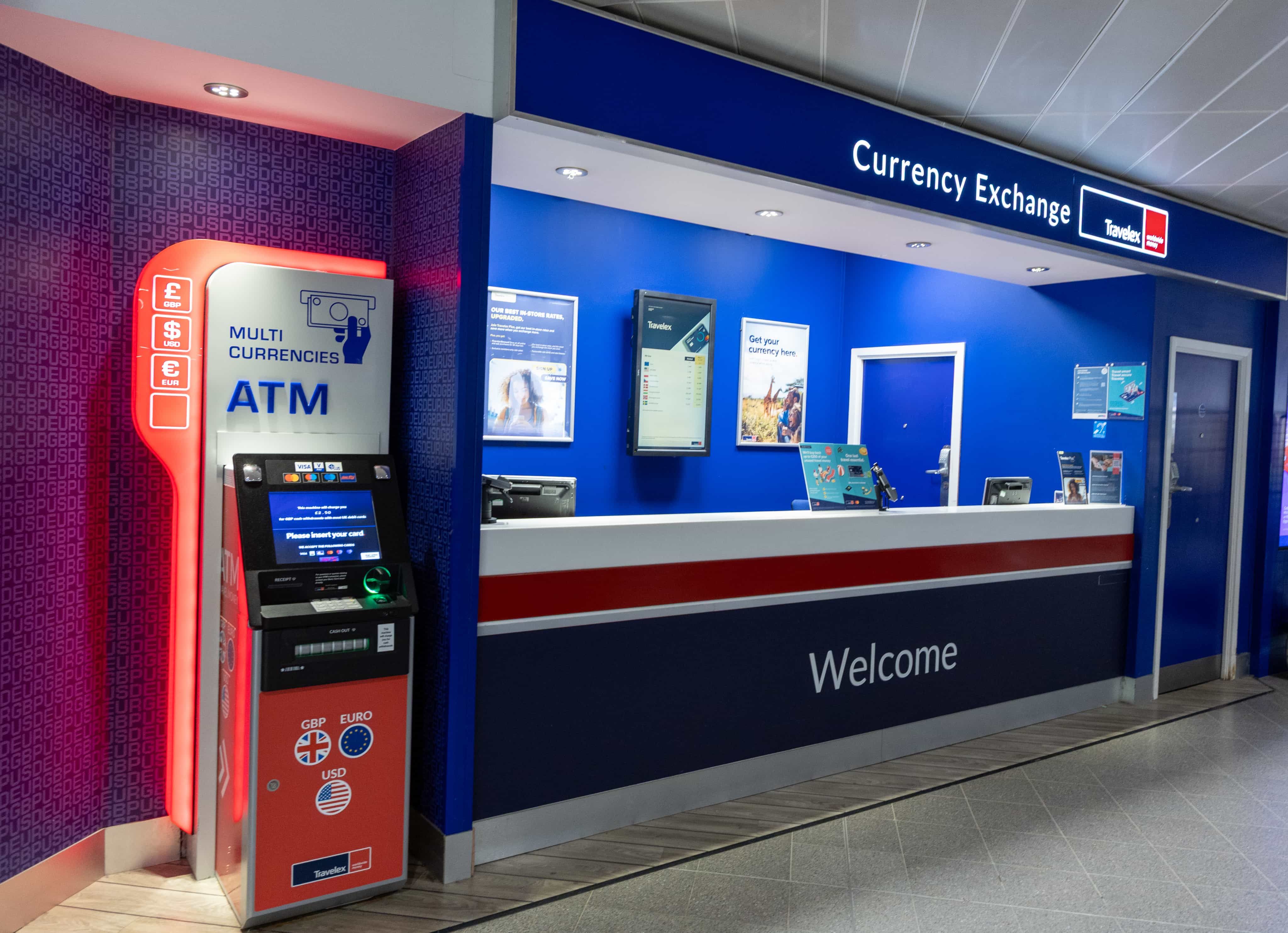 Currency exchange counter with a bright blue and white design, featuring a multi-currency ATM and welcome signage in an airport setting.