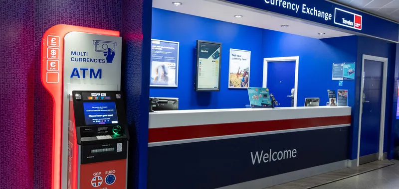 Currency exchange counter with a bright blue and white design, featuring a multi-currency ATM and welcome signage in an airport setting.