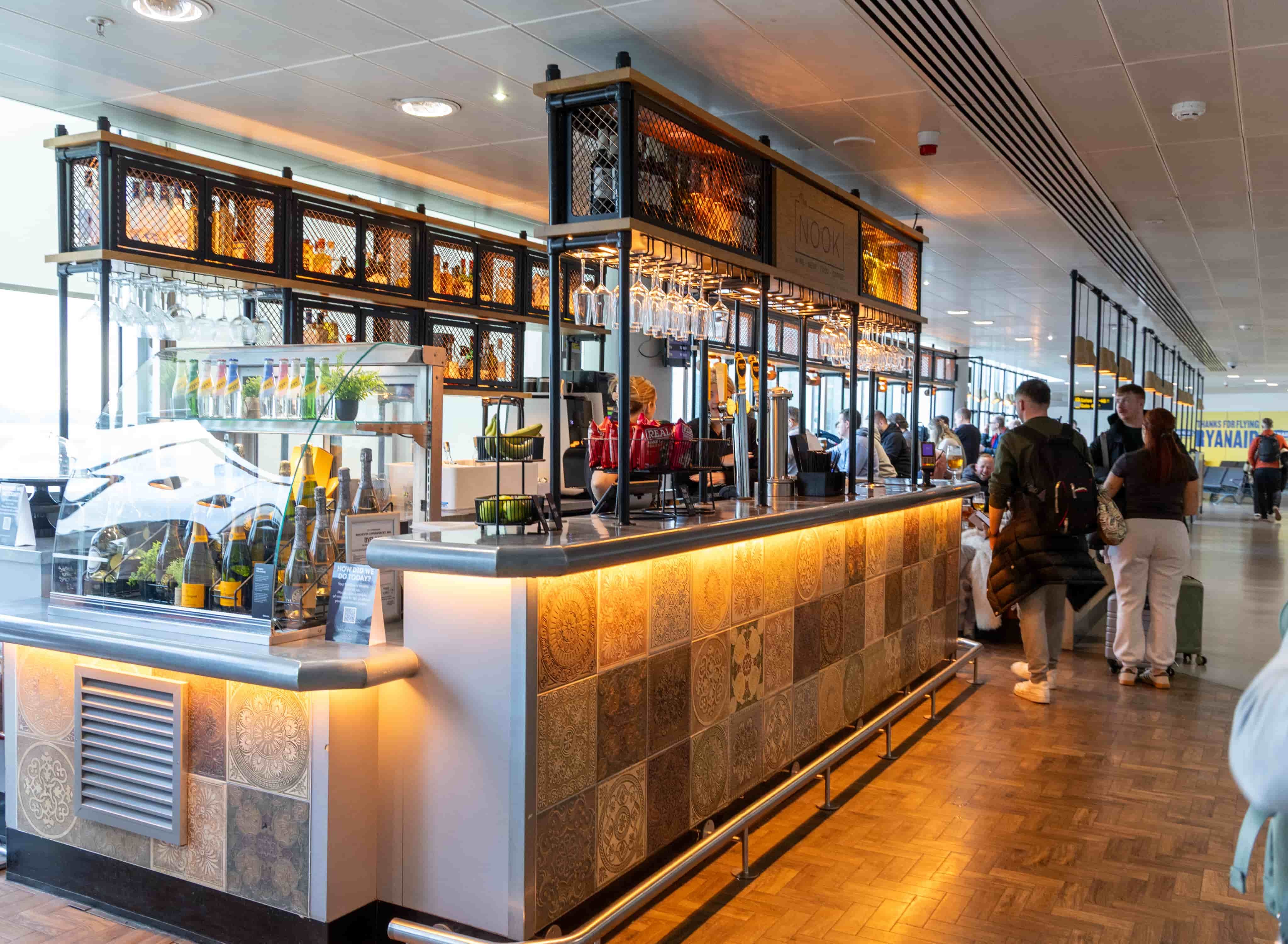 A modern airport café with a tile design, offering drinks and snacks. Passengers with luggage are passing by.