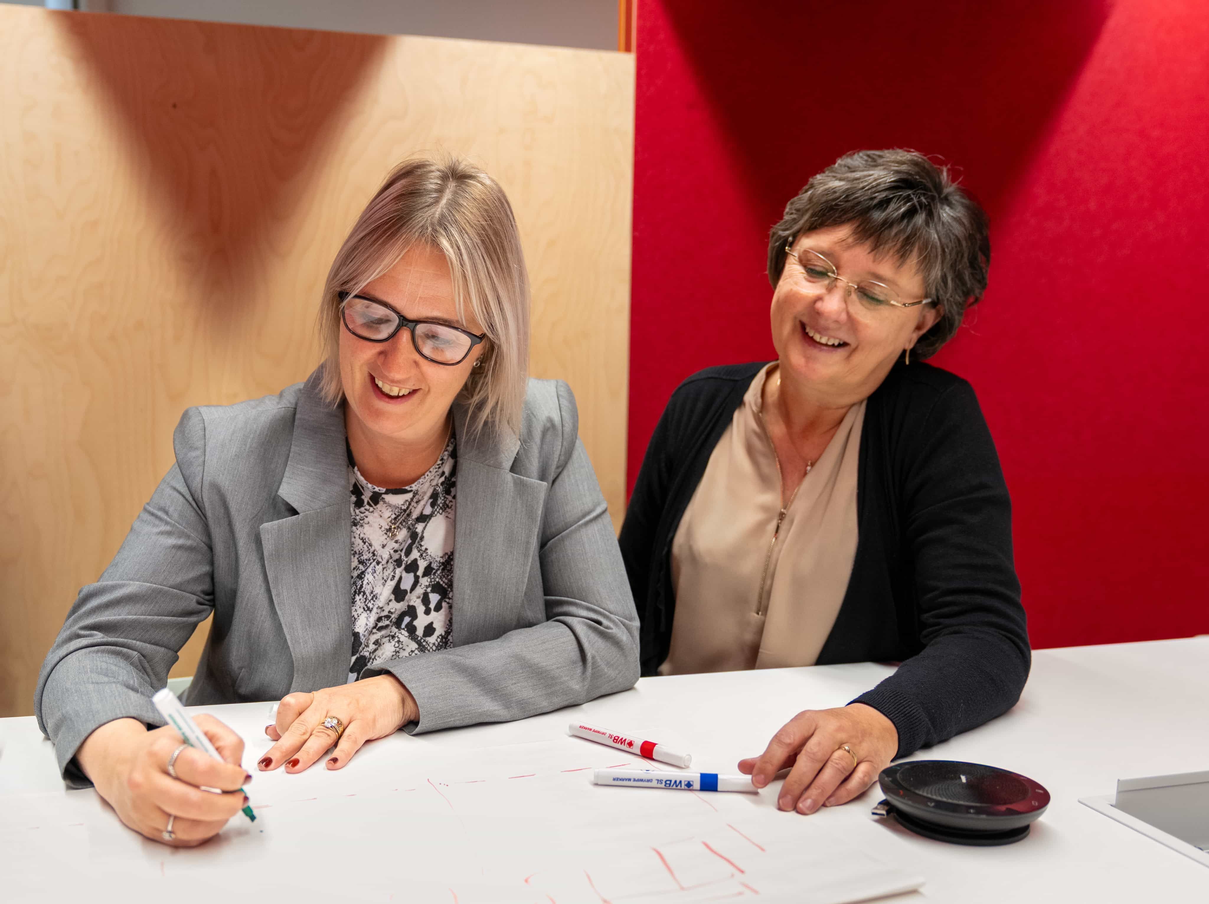 Two women smiling and collaborating at a table, one writing with a marker. Papers and additional markers are visible.