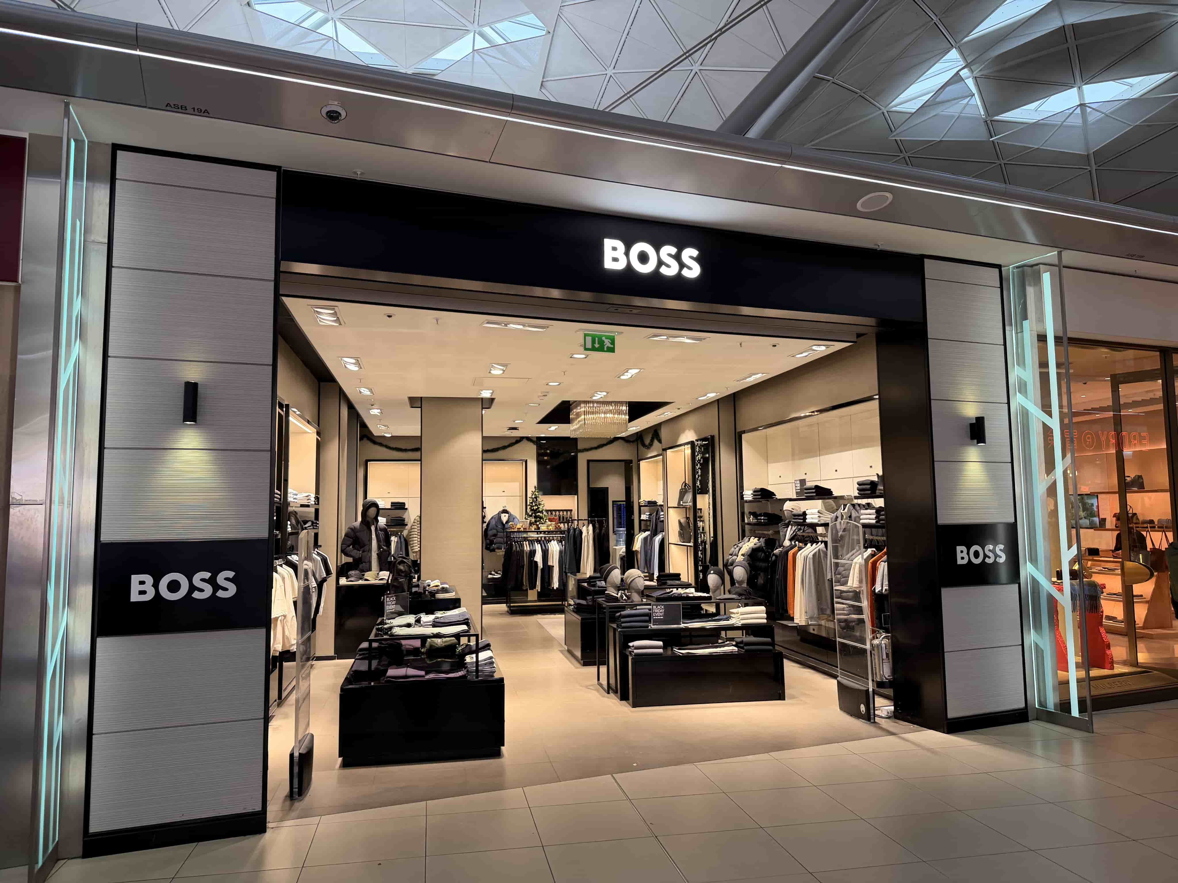 BOSS clothing store entrance in a mall, illuminated black sign, open interior with racks, tables and mannequins.
