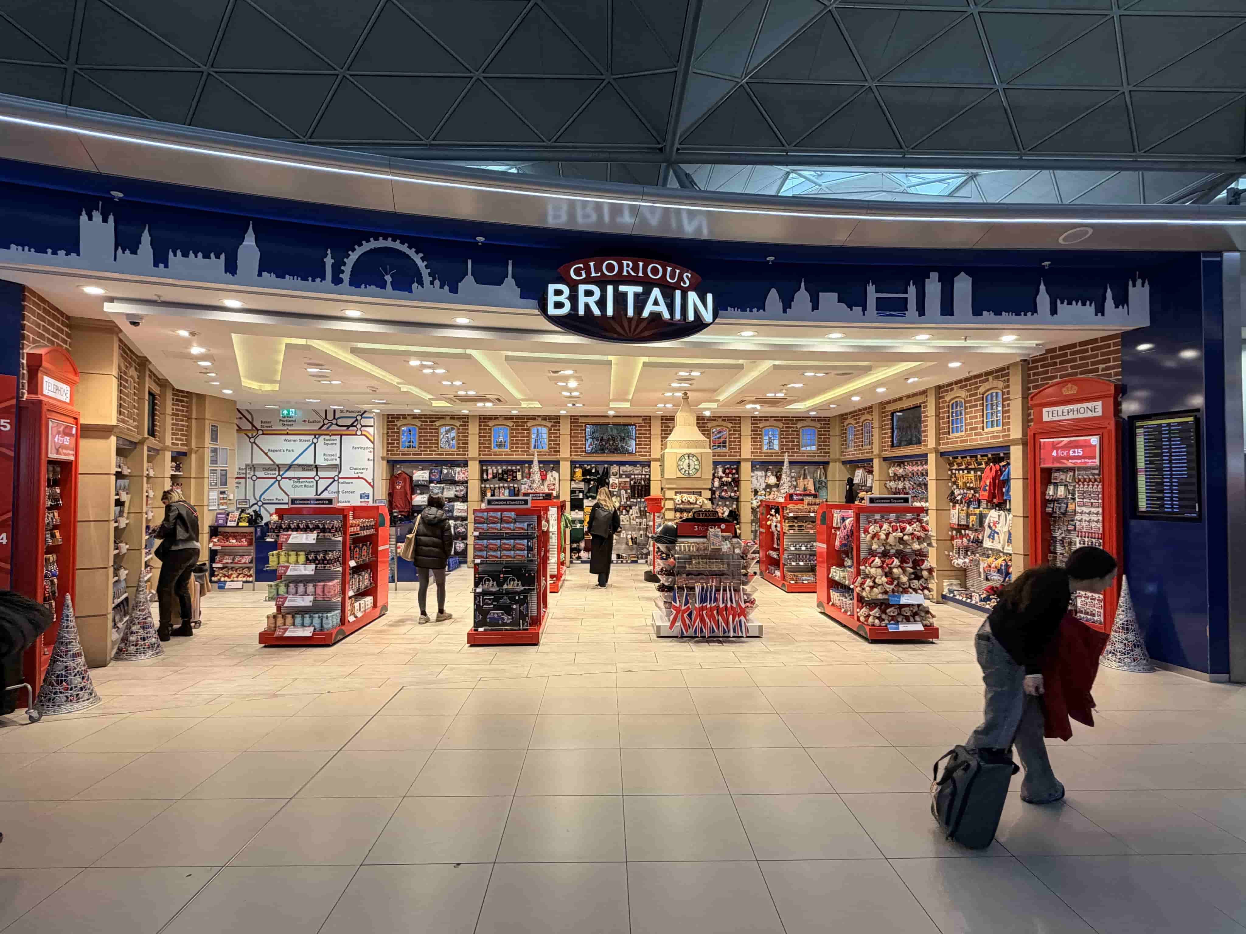 Interior view of 'Glorious Britain' souvenir shop with red displays, phone boxes and Union Jack items in an airport concourse.