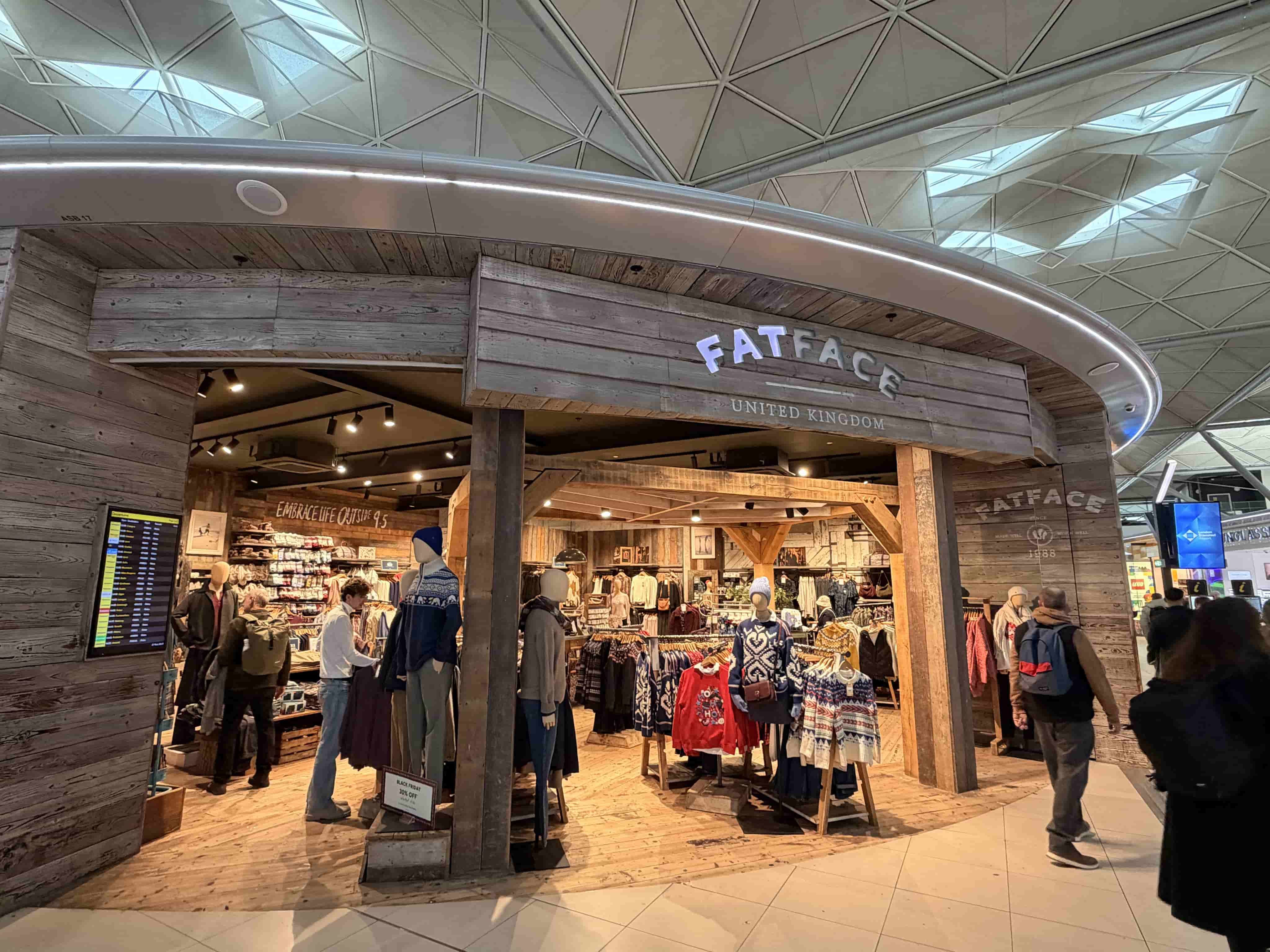 Rustic FatFace clothing store inside an airport terminal, wooden facade, mannequins and shoppers browsing.