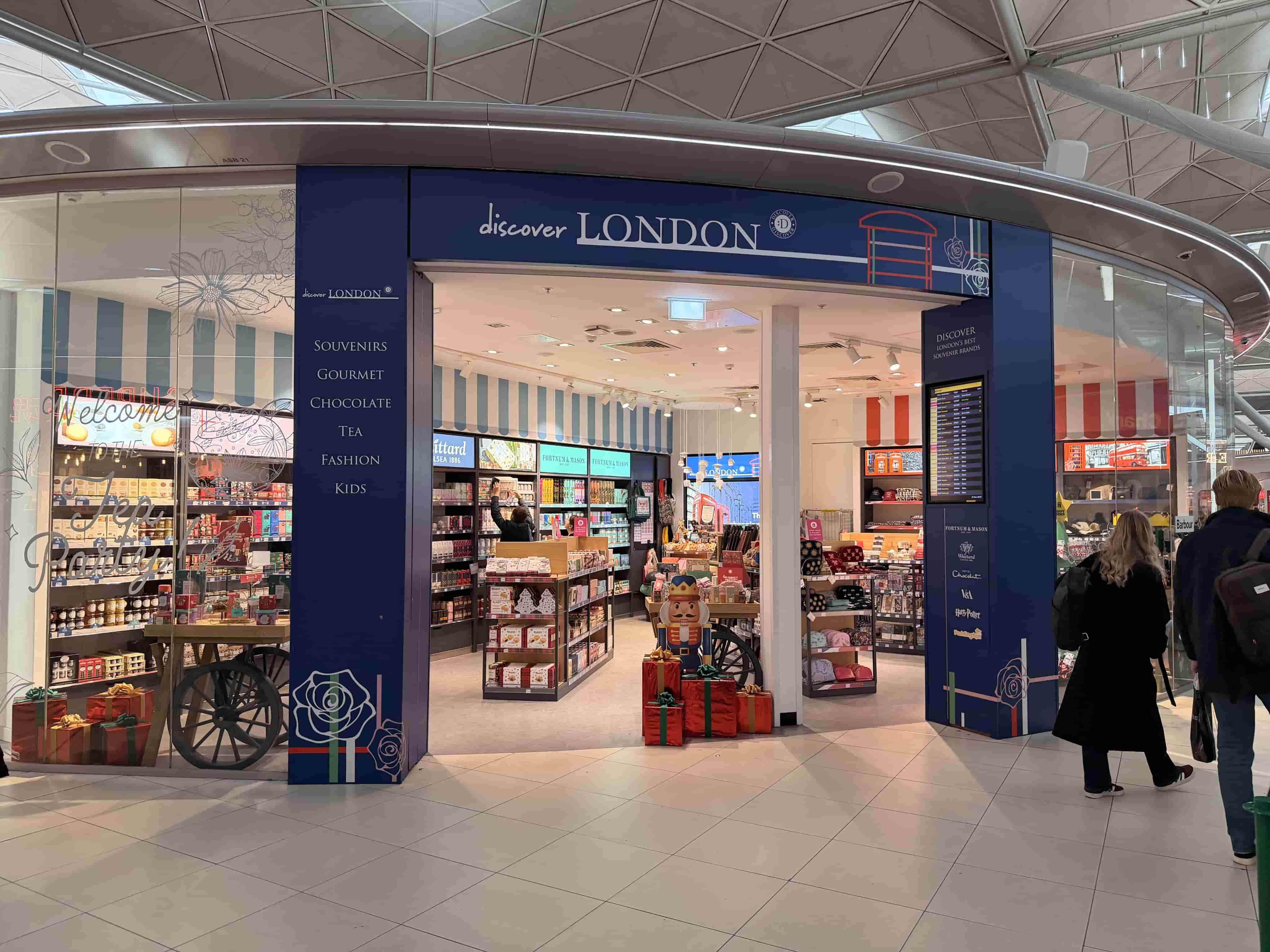 Airport storefront "discover LONDON" gift shop with blue facade, shelves of souvenirs, a nutcracker and wrapped presents, shoppers walking past.