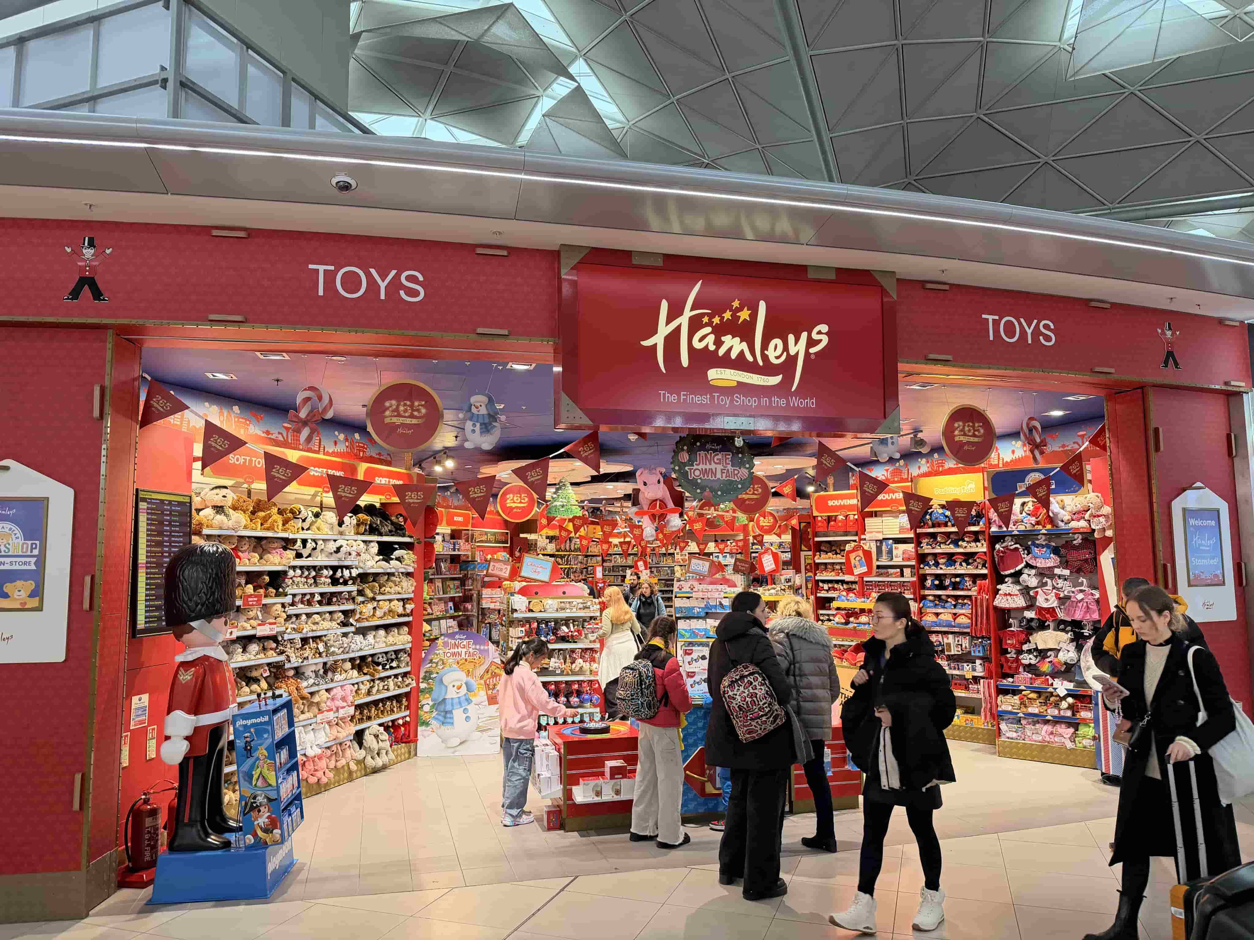Entrance of Hamleys toy store with red signage, giant toy soldier, shelves of plush toys and shoppers browsing the displays.