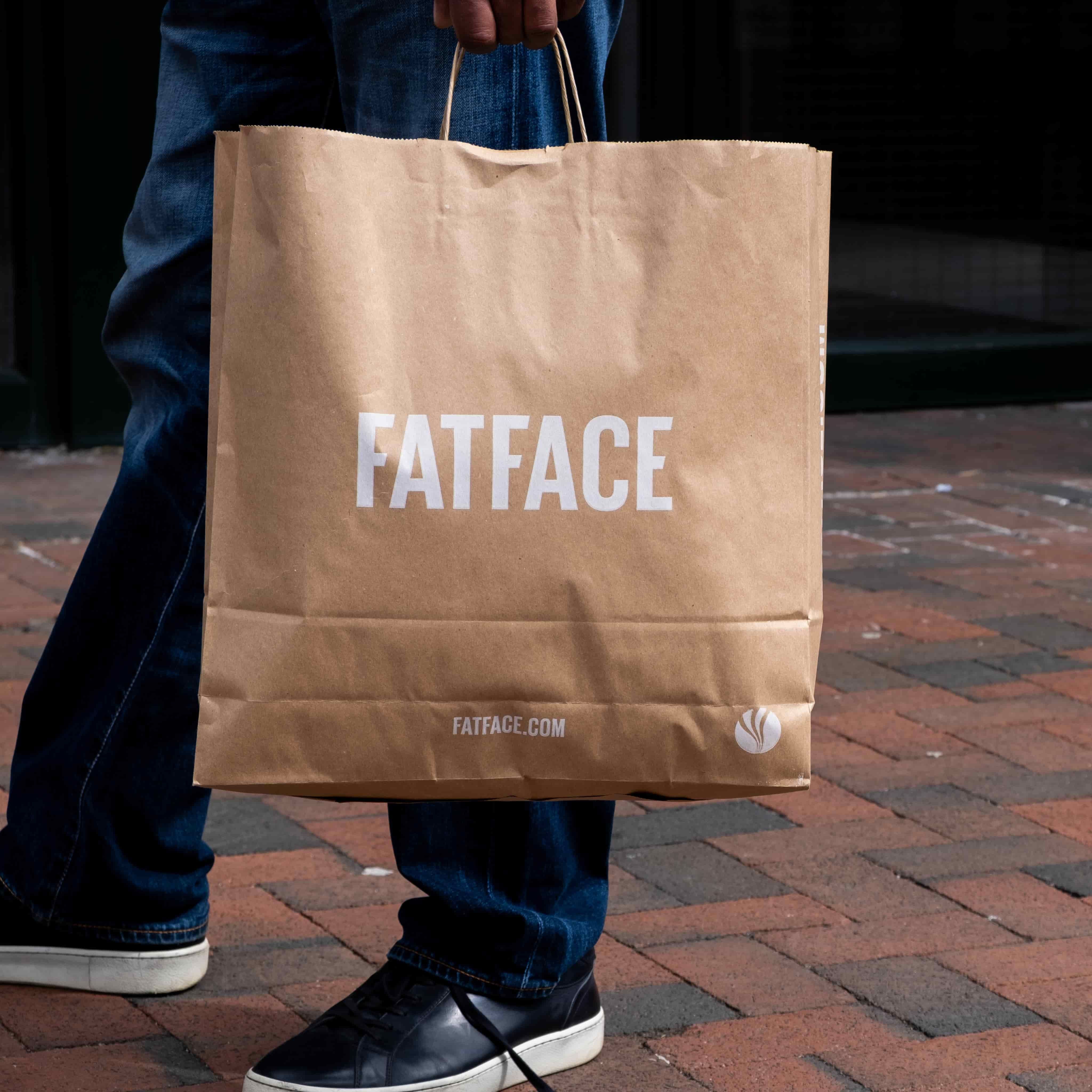 Hand holding a brown FATFACE paper shopping bag; person wearing blue jeans and black sneakers standing on brick pavement.
