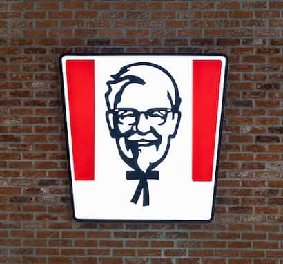 KFC logo featuring a stylized image of a man with glasses and a bowtie, set against a red and white background on a brick wall.
