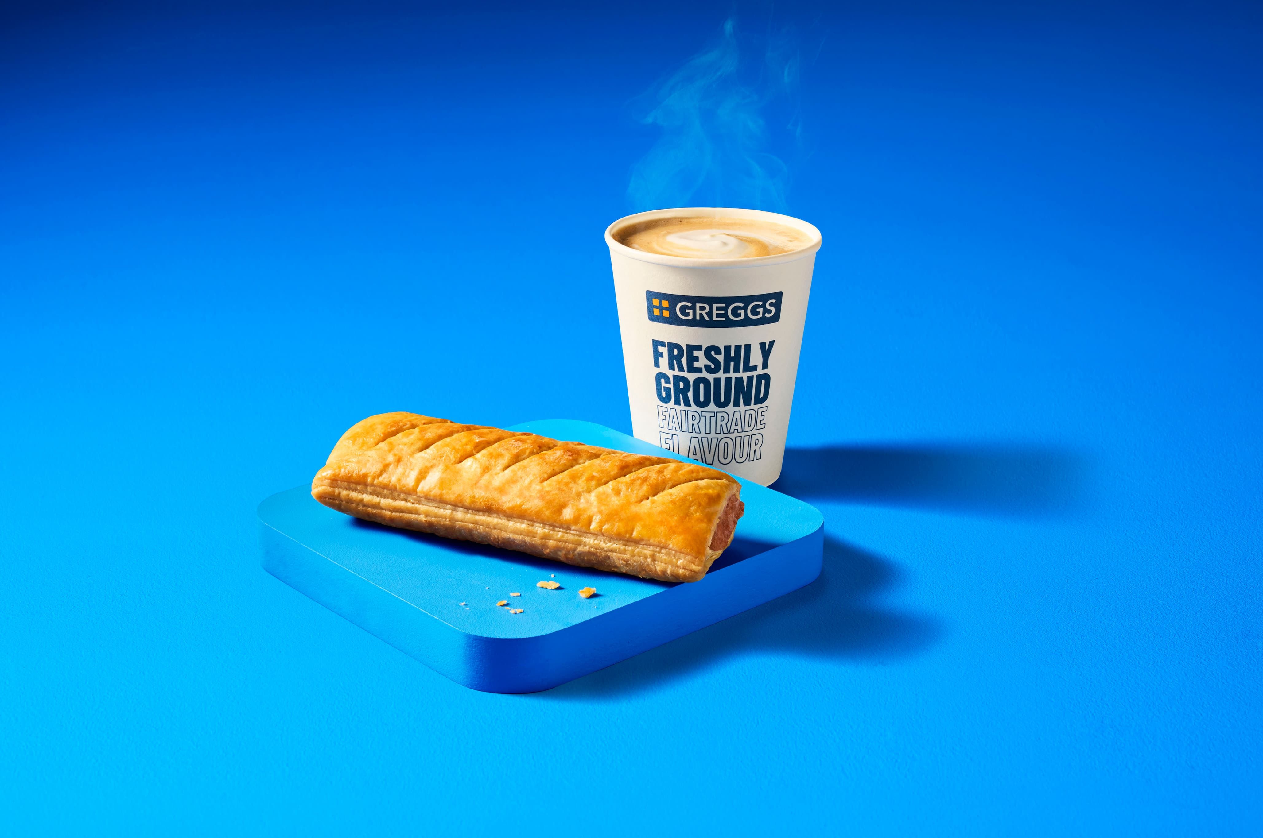 A steaming cup of coffee labeled "Greggs" next to a flaky pastry placed on a blue surface against a matching blue background.