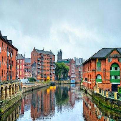 A canal lined with historic red brick buildings under a cloudy sky, reflecting in the water. A church tower is visible in the background.