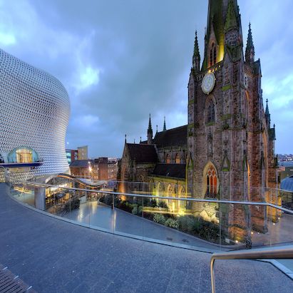 Modern building with a curved facade next to a historic church at dusk, featuring a glass walkway in the foreground.