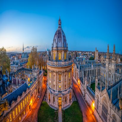 Aerial view of the Radcliffe Camera in Oxford at dusk, surrounded by historic buildings and softly lit streets under a clear sky.