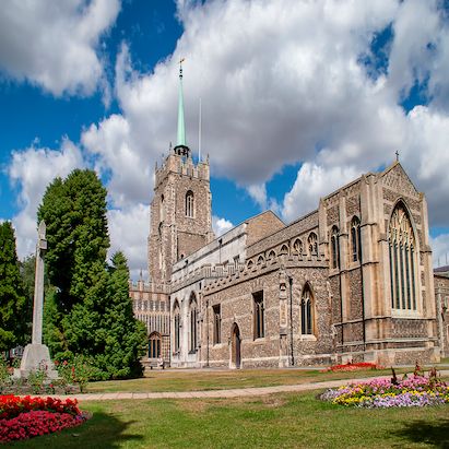 Historic stone church with a tall spire, surrounded by green lawns, colorful flower beds, and trees under a partly cloudy sky.