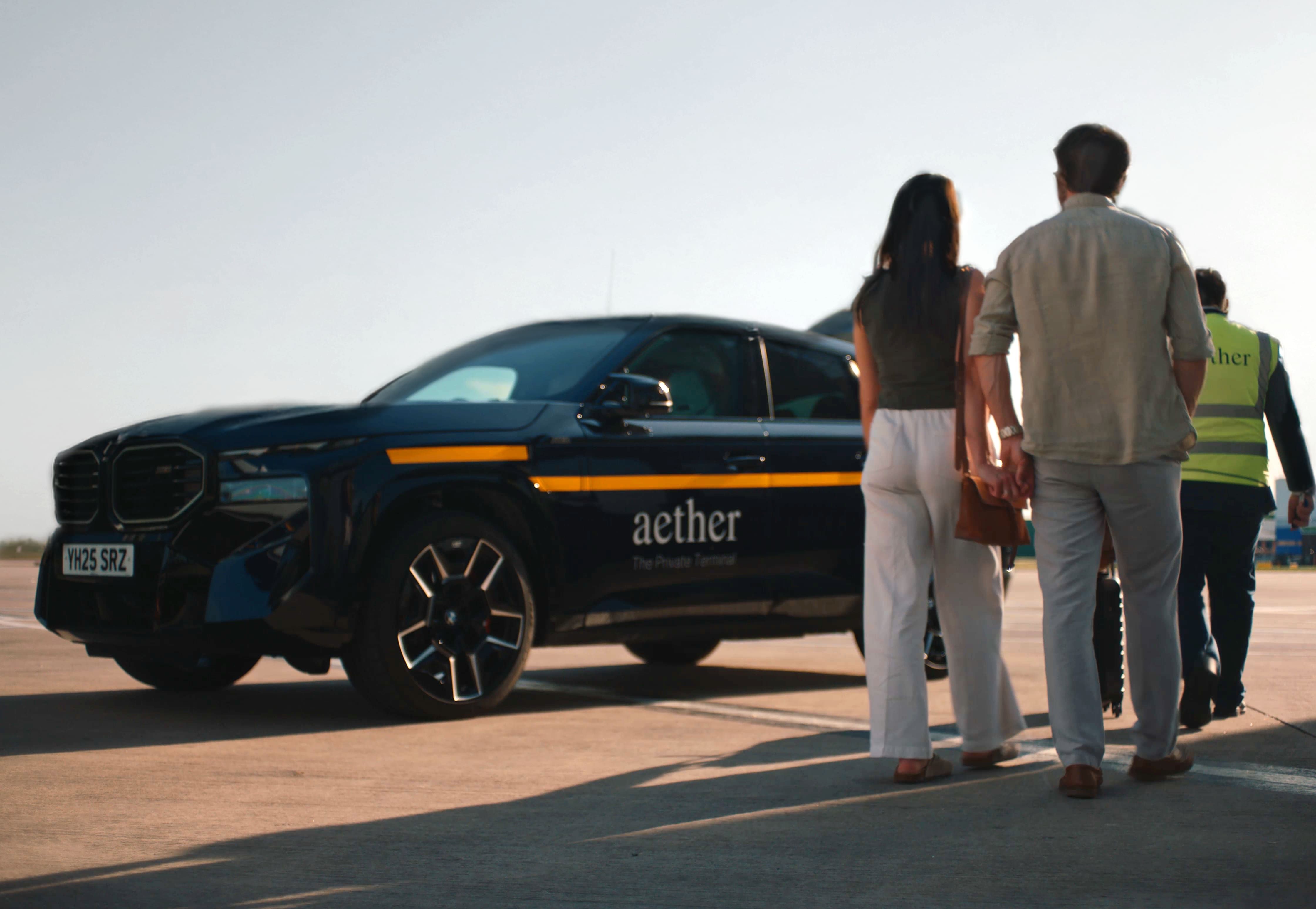 A couple walks towards a black car with an "aether" logo, accompanied by a person in a reflective vest on a sunny day.