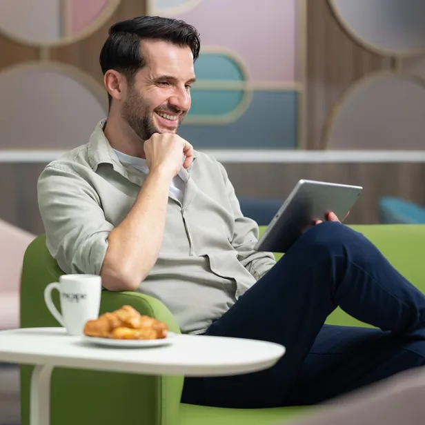 A man in a casual shirt sits on a green chair, smiling while holding a tablet. A plate of croissants and a mug are on the table in front of him.