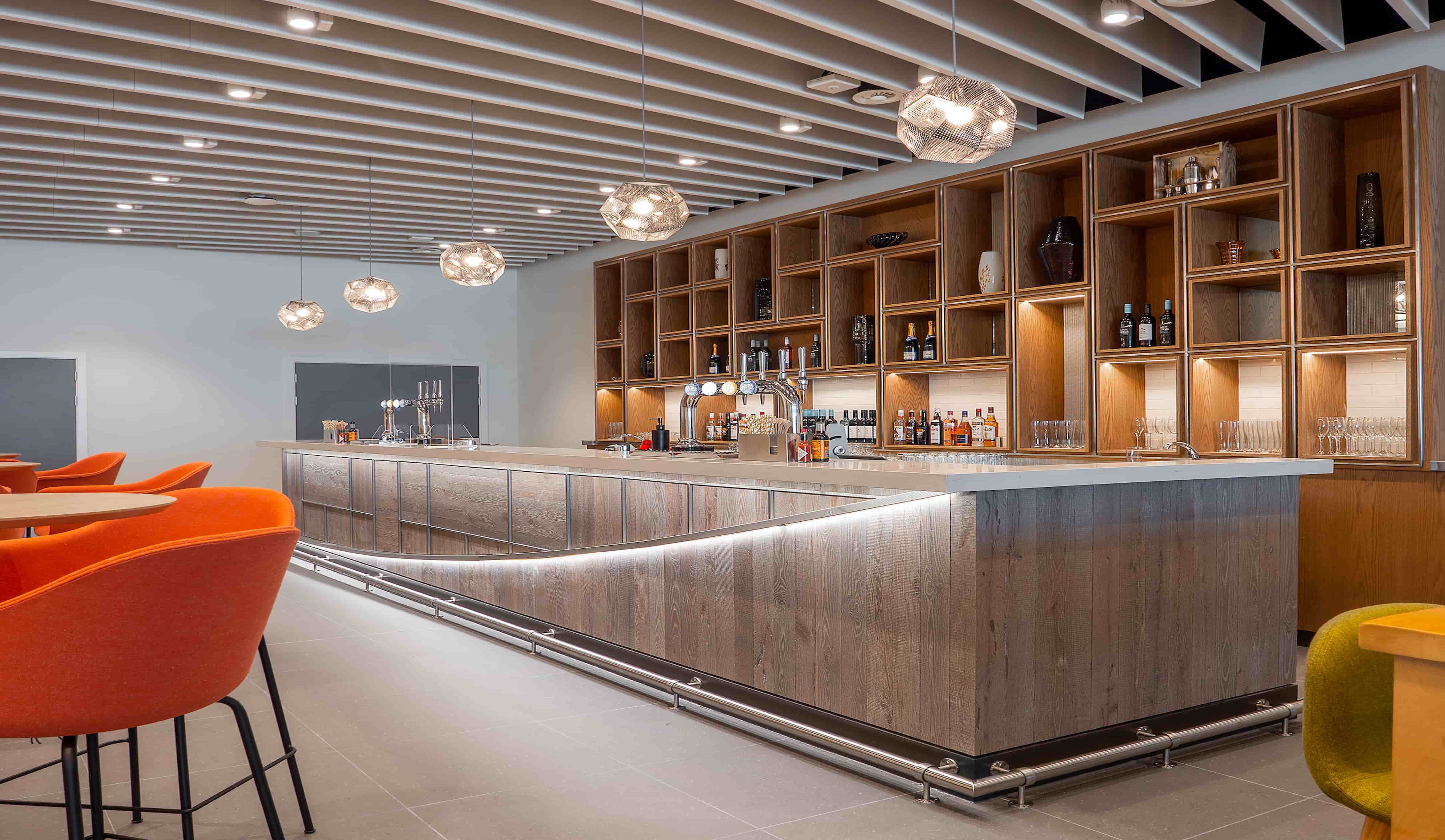 Modern bar interior with wooden shelves stocked with bottles, pendant lights hanging above a long counter, and orange chairs in the foreground.
