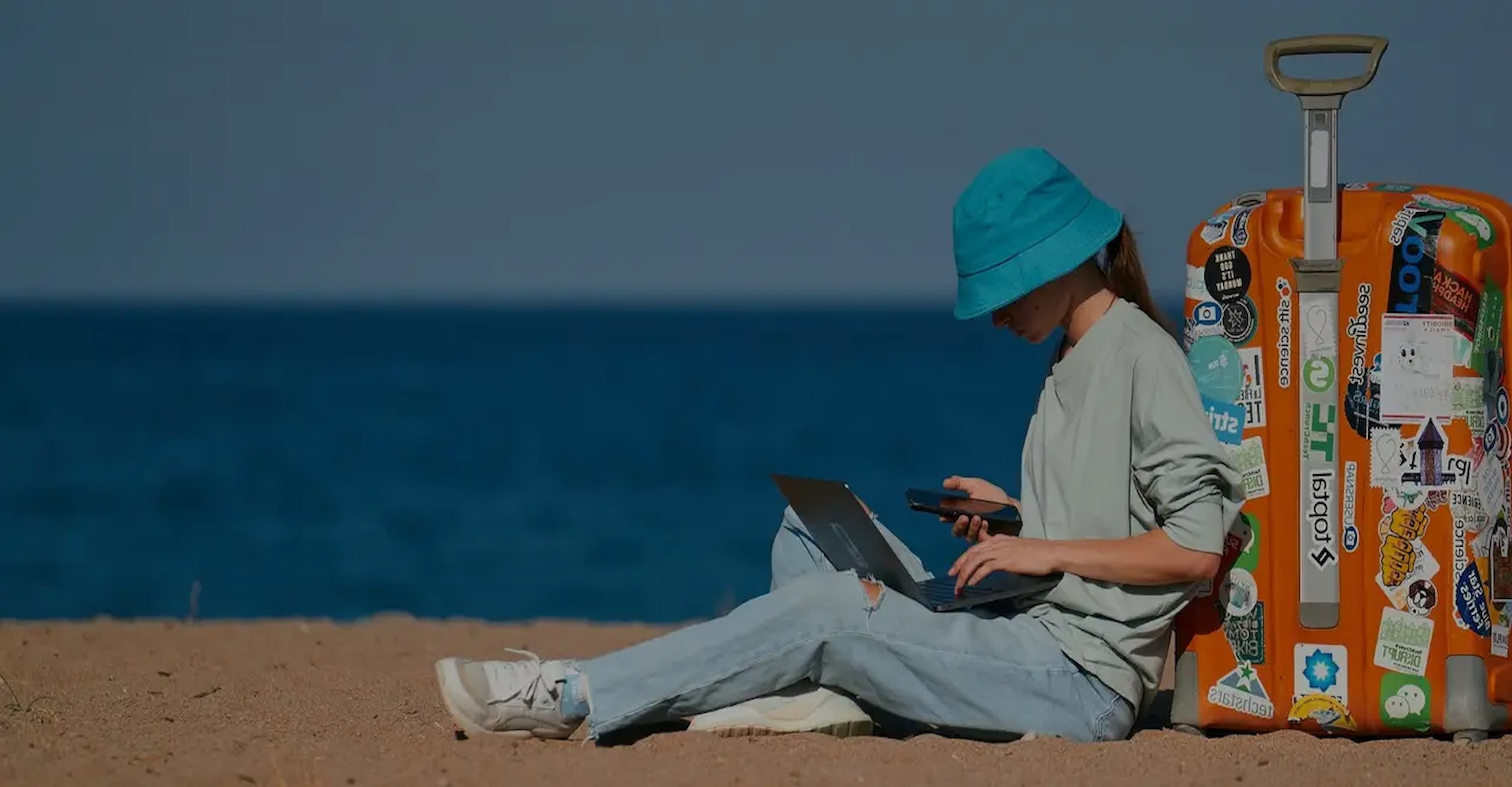 Person in a blue hat sitting on a beach using a phone and laptop, with a sticker-covered orange suitcase nearby.