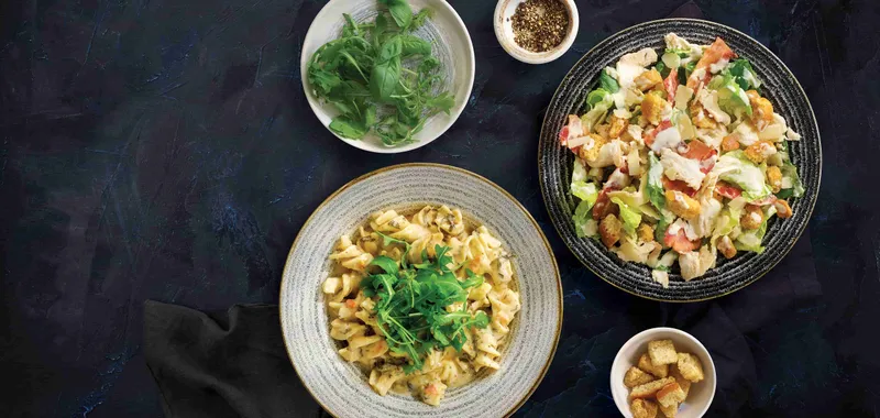 A plate of creamy pasta topped with greens, a bowl of mixed salad with croutons, a small bowl of spices, greens, and a cup of croutons on a dark background.
