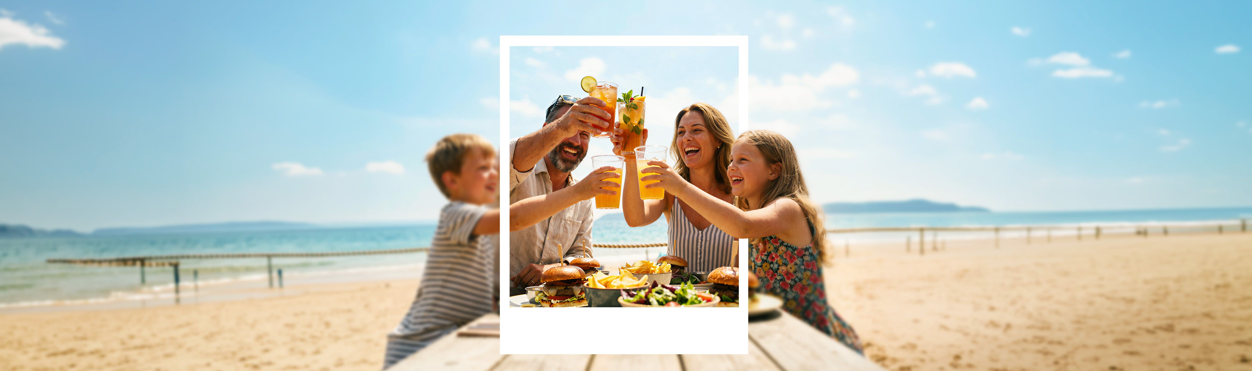 A family seated at a beachside table raising glasses in a celebratory toast, with sandwiches and salads on the table and the ocean in the background.
