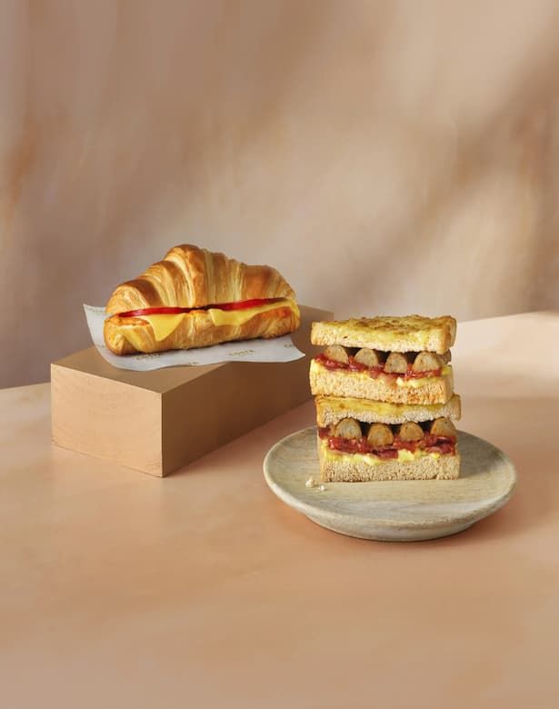 A croissant with cheese on a wood block and a plate with stacked grilled sandwiches filled with sausages and sauce.