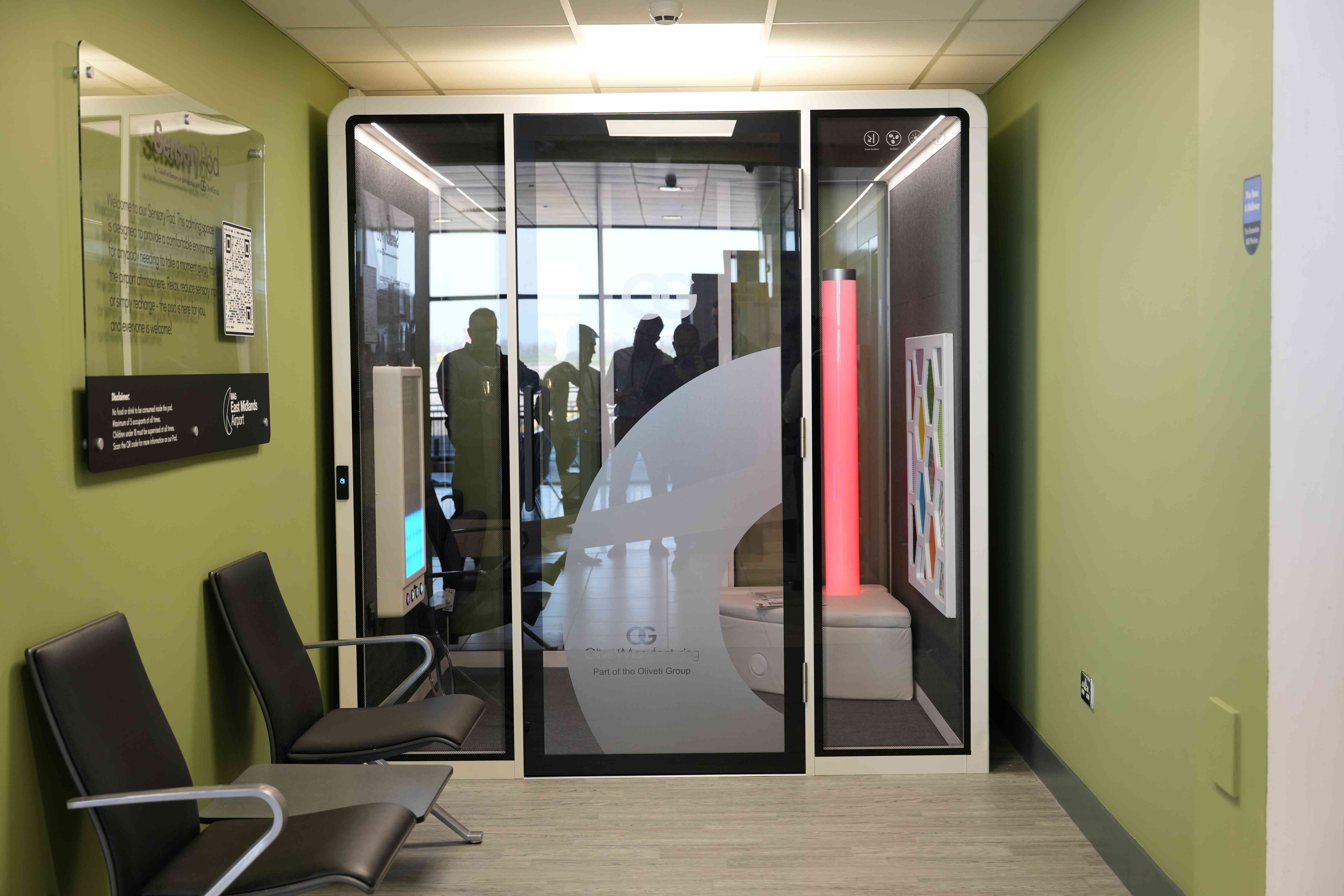 Modern office booth with glass door, surrounded by green walls. Two chairs are to the left. Silhouettes visible inside the booth.