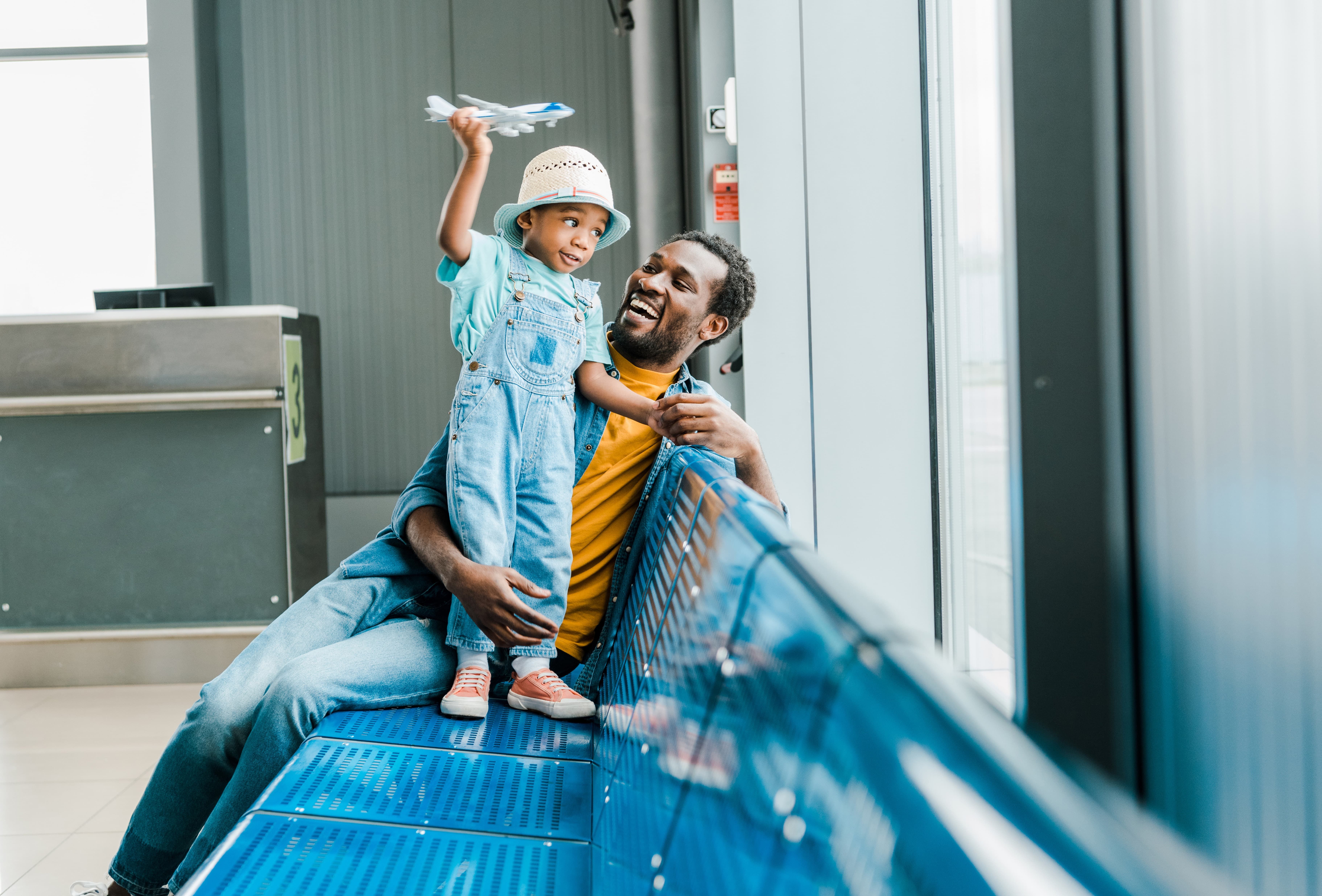 Man joyfully holds child with toy airplane on airport bench. Child wears a hat and overalls; both smile brightly near large windows.