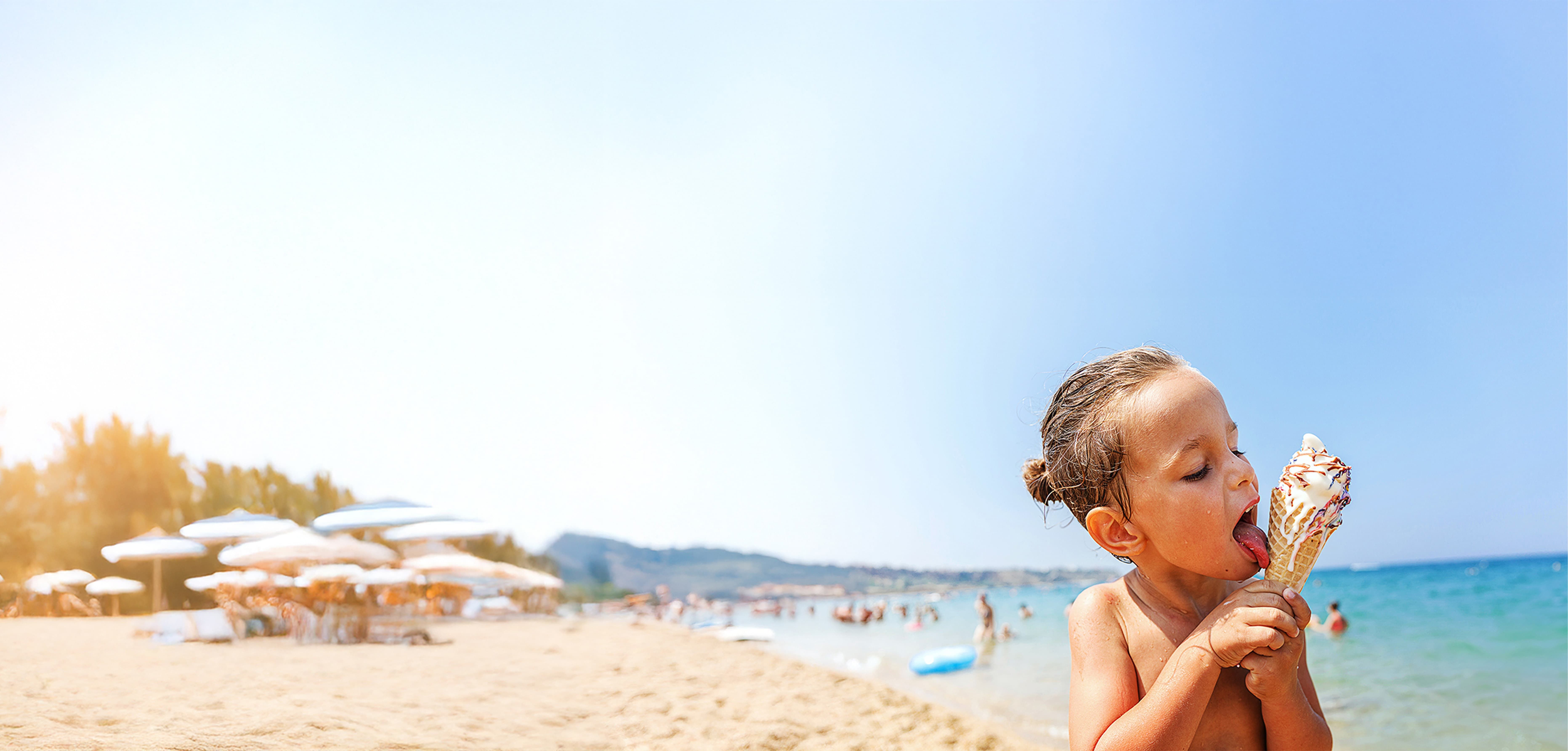 Child enjoying an ice cream cone on a sunny beach, with umbrellas and people in the background.