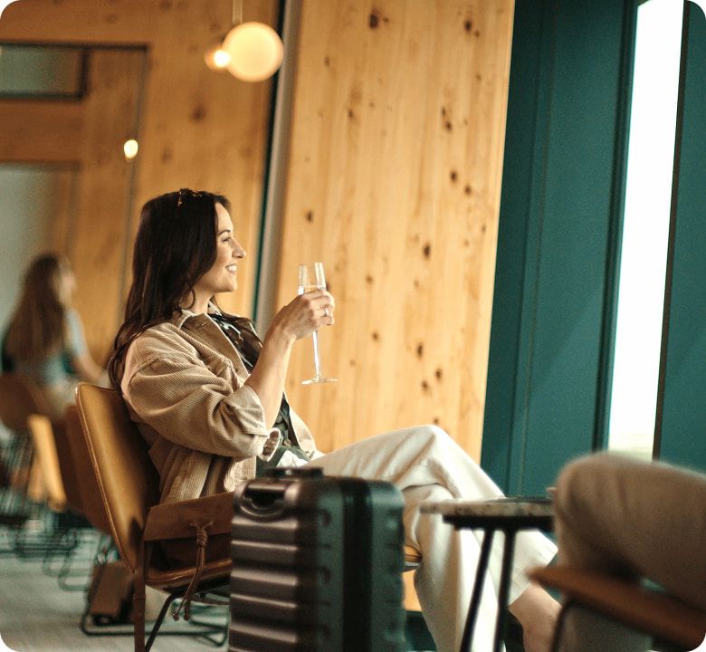 Woman sitting in an airport lounge, holding a glass, with luggage by her side, looking out the window. Warm, wooden interior decor.