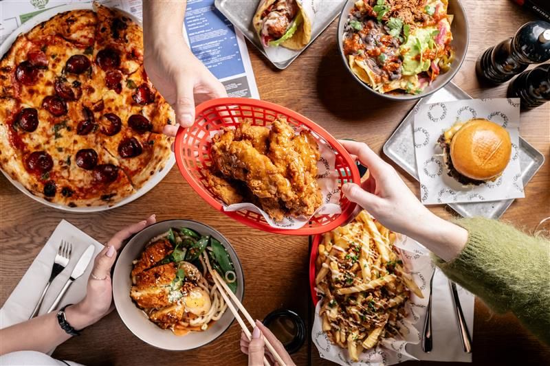 A variety of dishes on a table including pizza, fried chicken, salad, burger, loaded fries, and noodles, with people sharing and serving food.