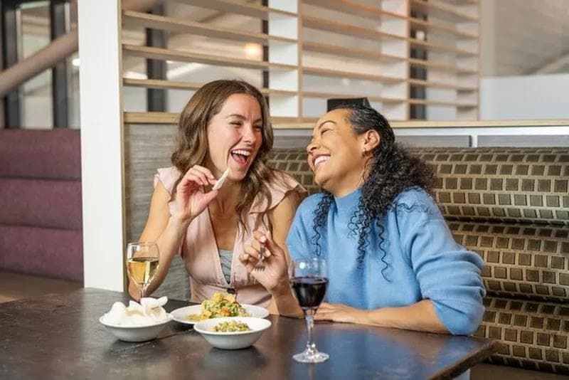 Two women sitting at a restaurant table, laughing and holding hands. They have glasses of wine and bowls of food in front of them.