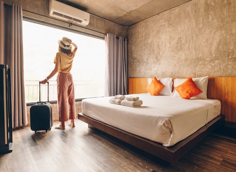 A person with a hat stands in a hotel room by a large window, holding a suitcase. The room has a bed with orange pillows and folded towels.