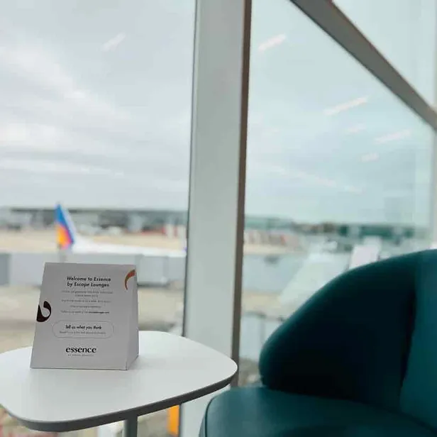 A lounge area with a sign reading "Welcome to Essence by Escape Lounges" on a small table, and an airport runway visible through the window.